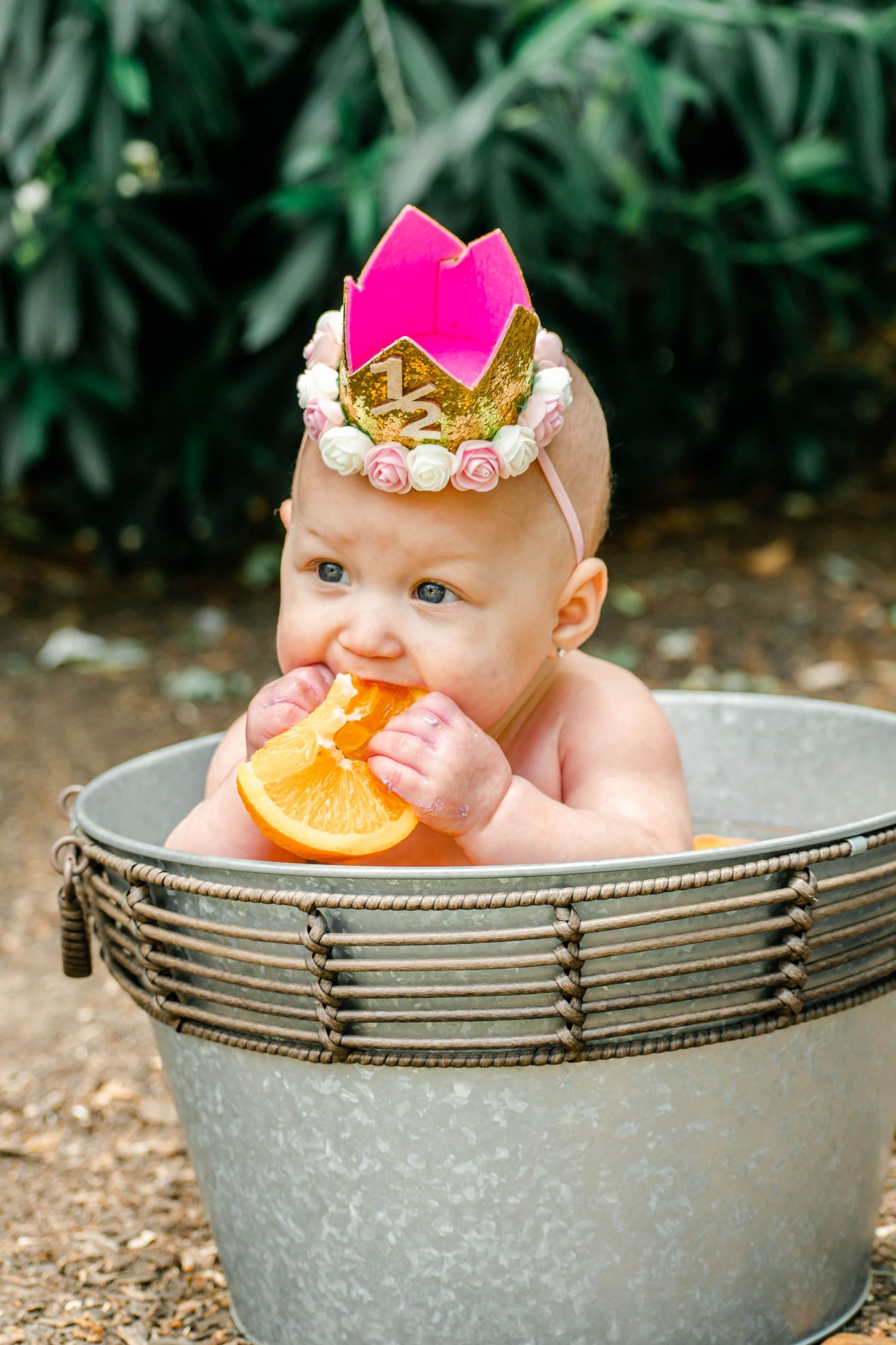 Baby with pink and gold crown explores orange slices during outdoor photoshoot in metal container.