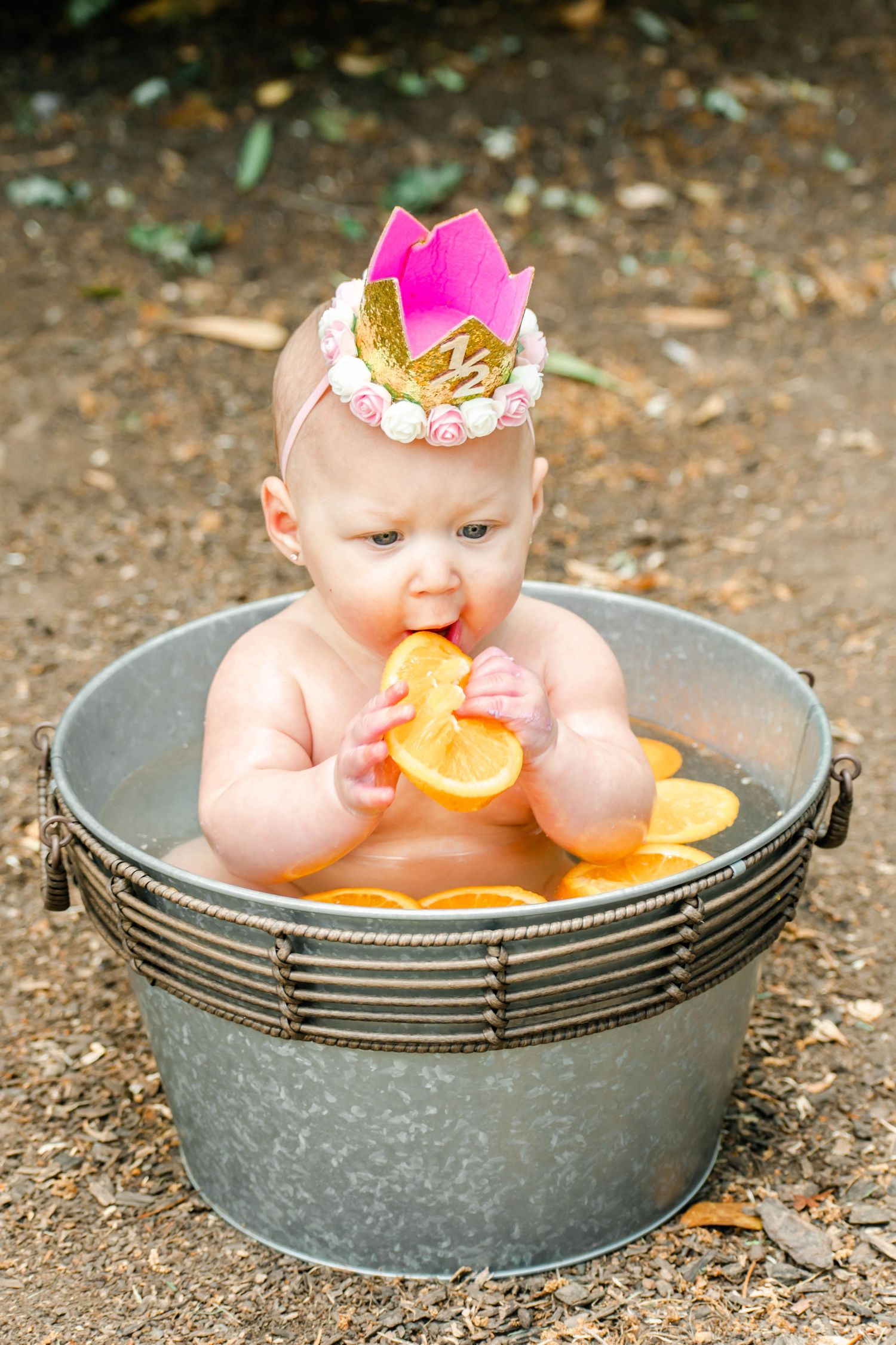 Infant wearing floral crown plays with orange segments while seated in vintage metal wash basin.