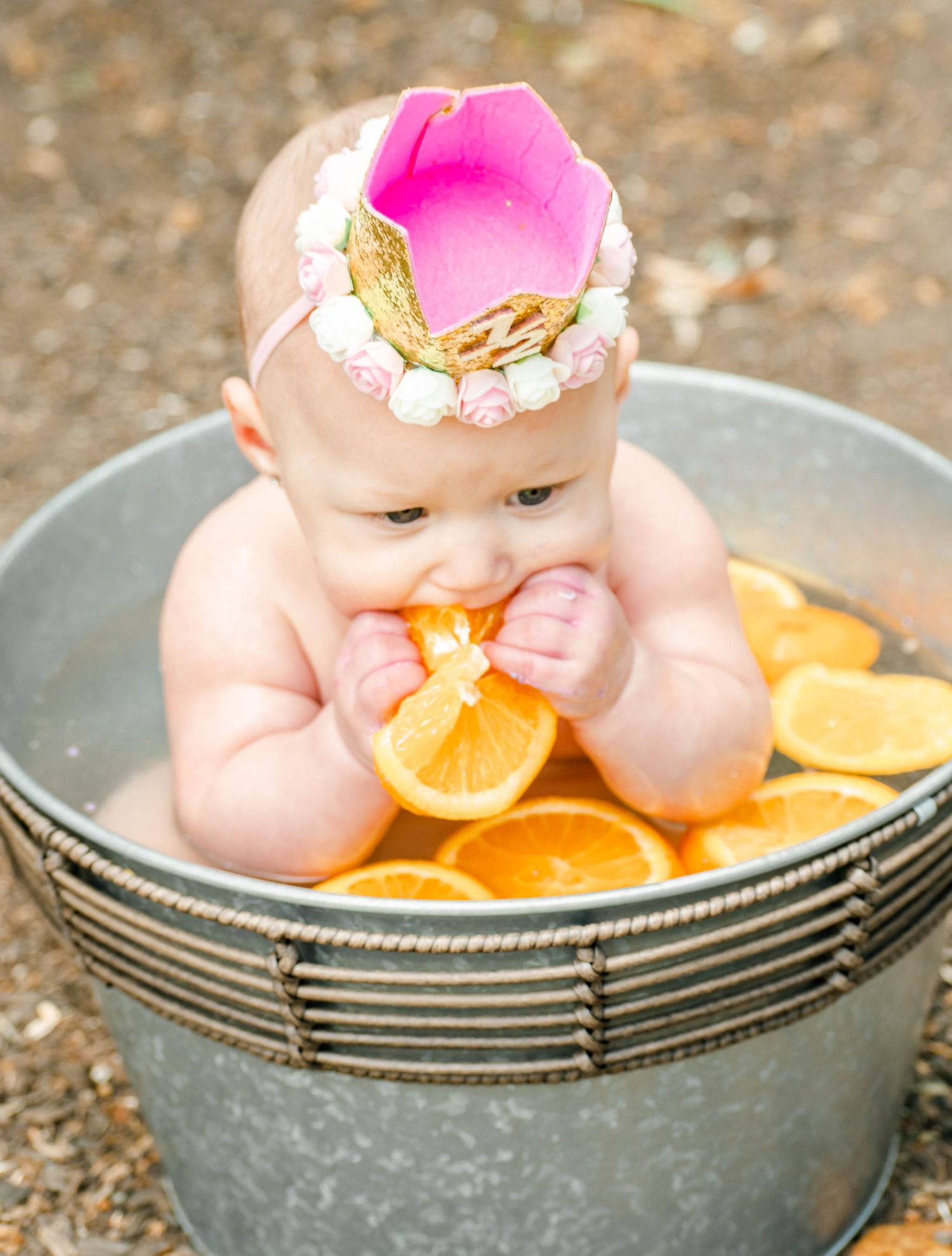 Creative baby photography setup with infant posed in metal tub with sliced oranges and pink accessories.