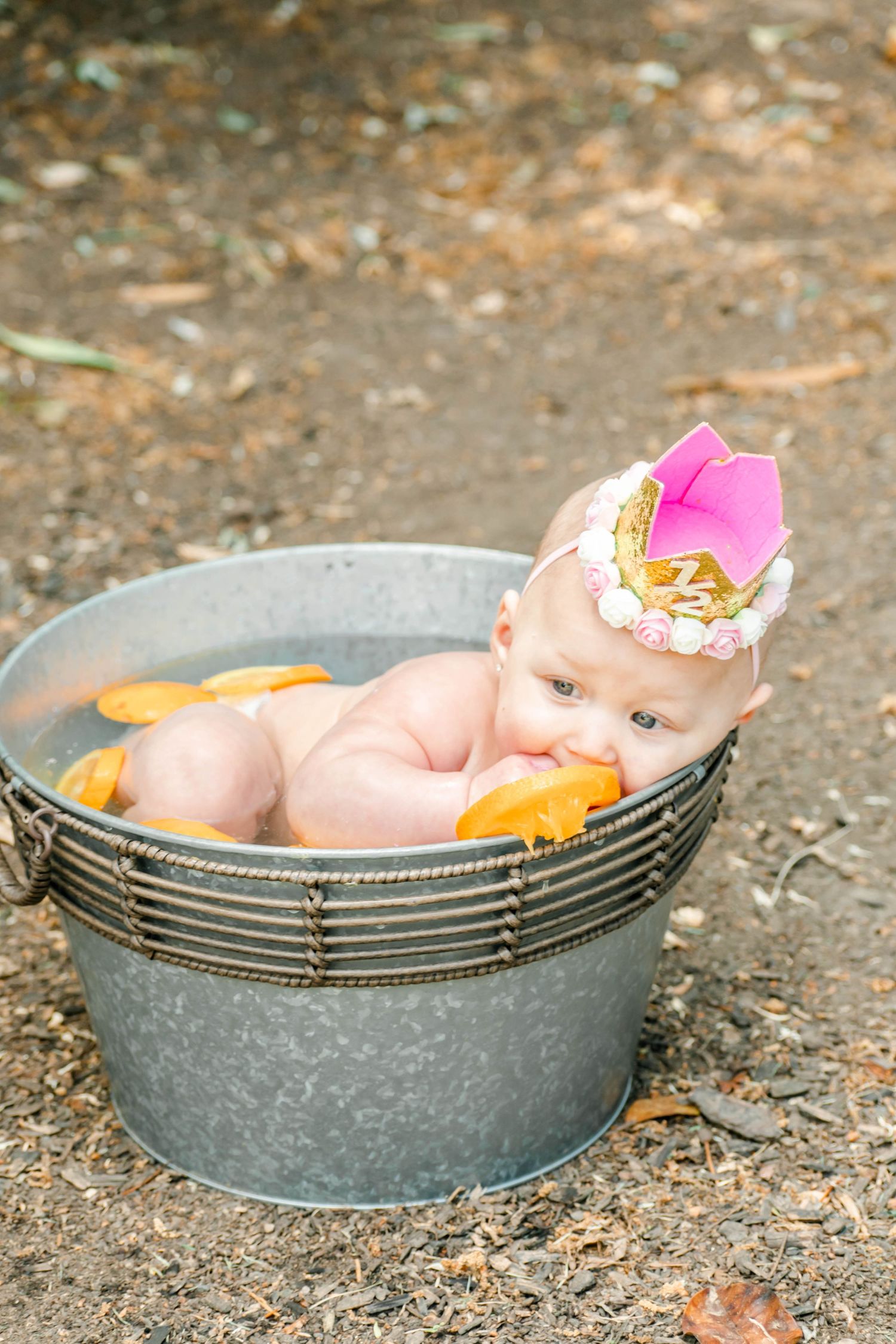 Baby in pink crown enjoys citrus fruit while posing in galvanized metal tub outdoors.
