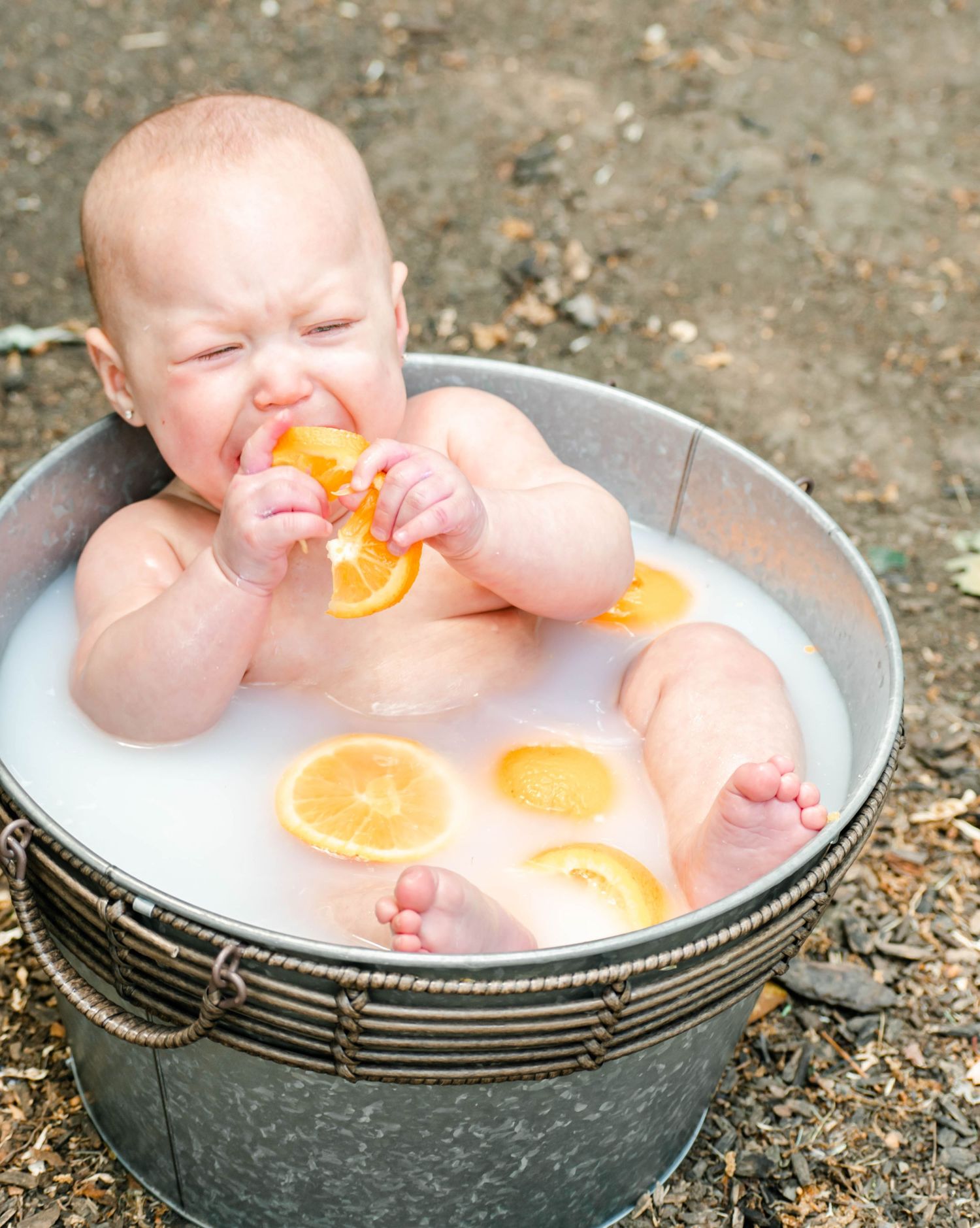 A baby in a milk bath with orange citrus slices floating around playfully interacts with the fruit.