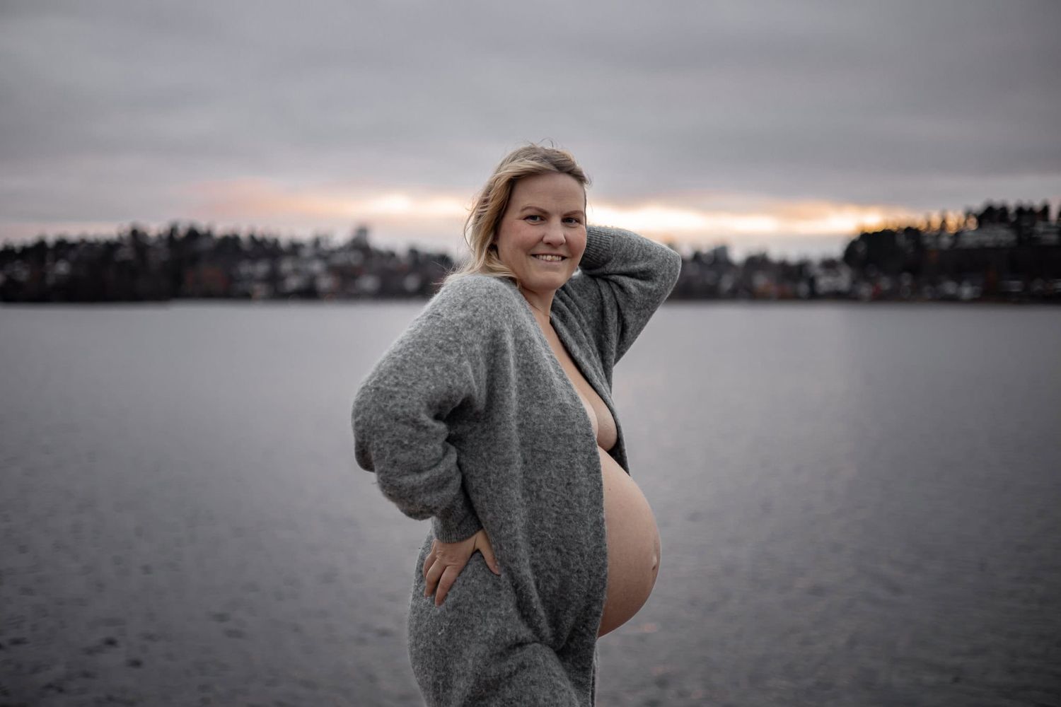 Gravid kvinna med kofta och bar mage i en gravidfotografering utomhus. Solnedgång och moln bakom, hon ser in i kameran och ler.
