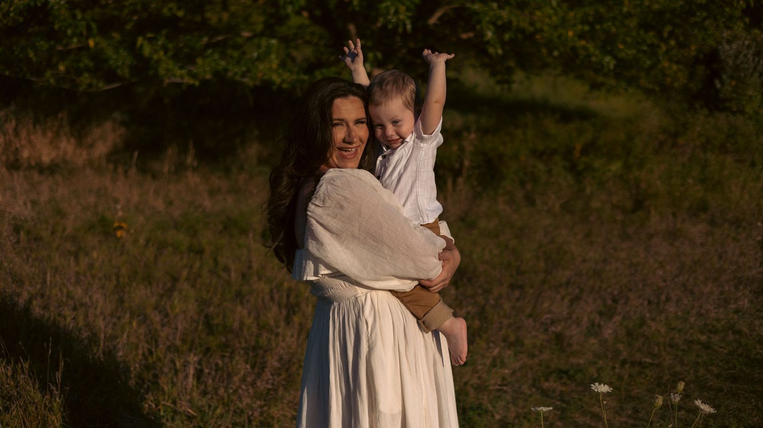 A couple embraces in a rustic outdoor setting during golden hour with tall grass and trees in the background.