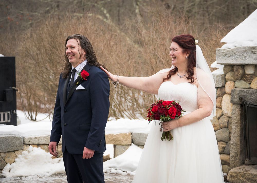 Winter wedding couple poses in front of snow-covered stone wall with red floral accents.