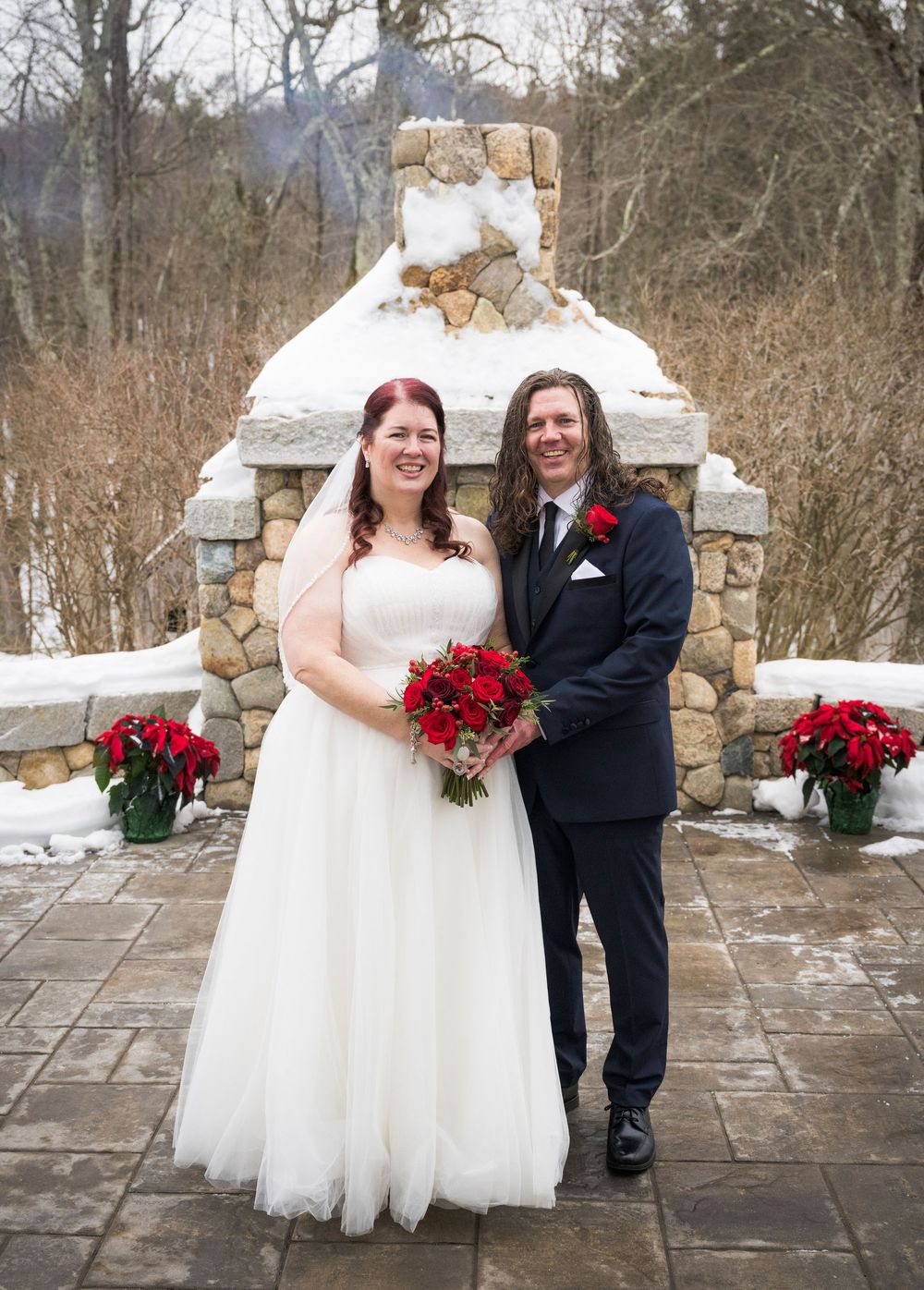 Newlyweds share joyful moments on a stone patio with red bouquets against a winter landscape.