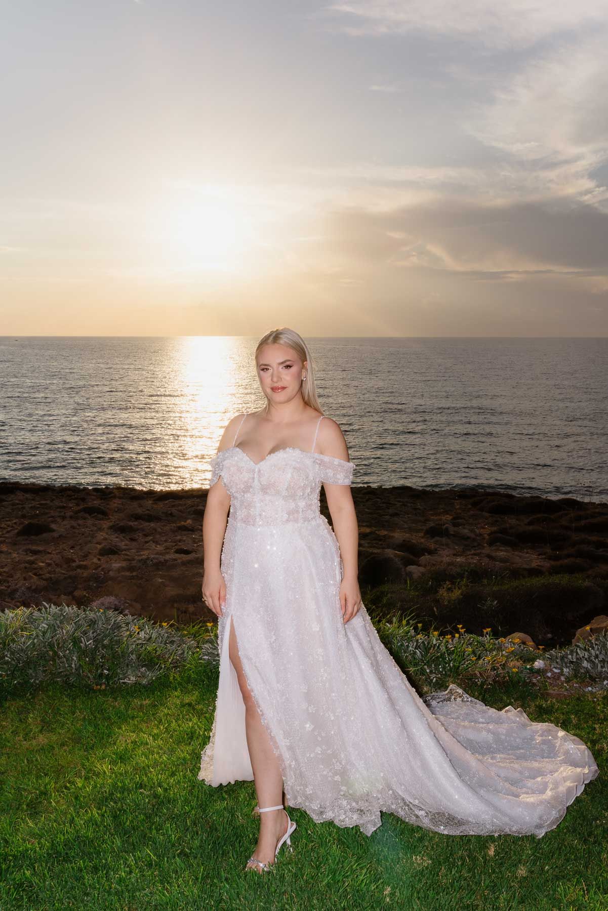 A sunset photoshoot by the ocean features someone in a flowing white lace dress reclining on coastal grass.