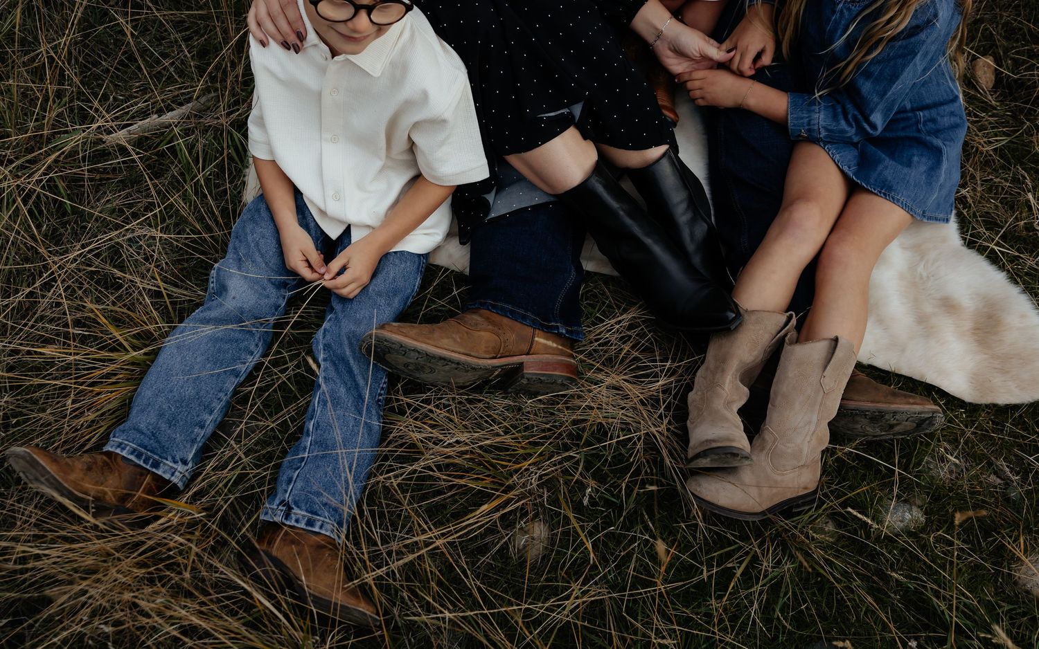 People in casual denim and boots sitting on grass with a cozy blanket during an outdoor gathering.