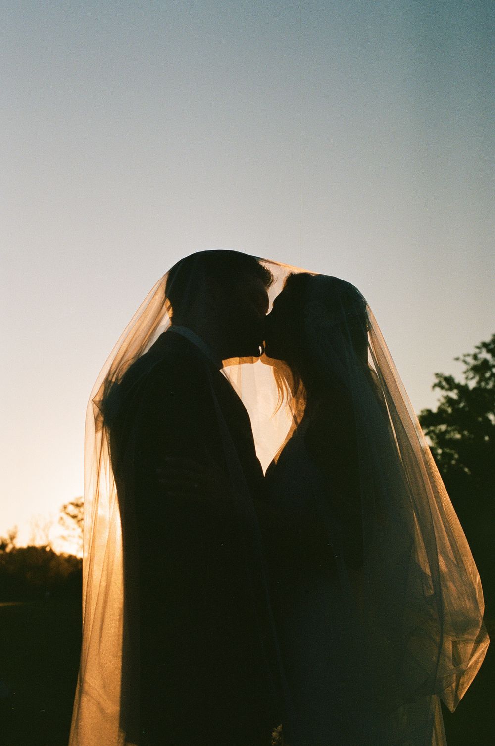 Silhouettes of a couple against sunset sky with backlit veil creating romantic atmosphere.