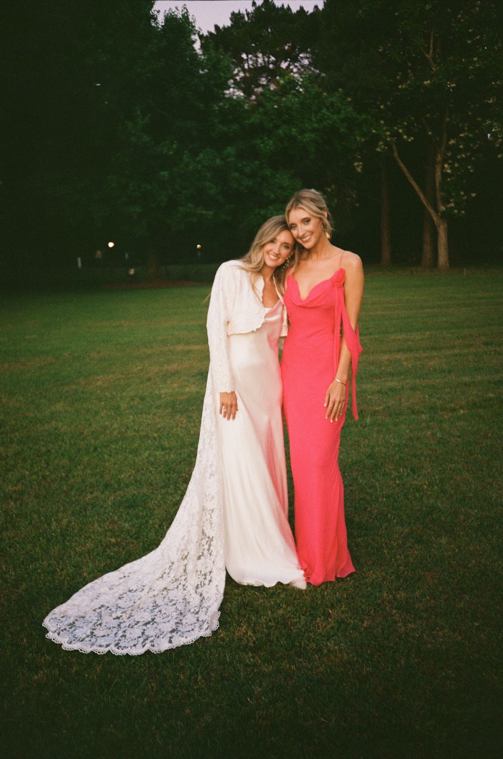 Two people in formal dresses, one in white lace with a train and one in coral pink, stand together on a grassy lawn at dusk.