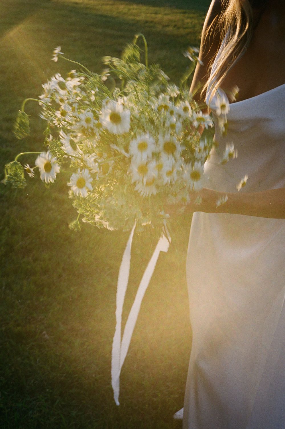 A delicate bouquet of white daisies and wildflowers held against a flowing white wedding dress in sunlight.