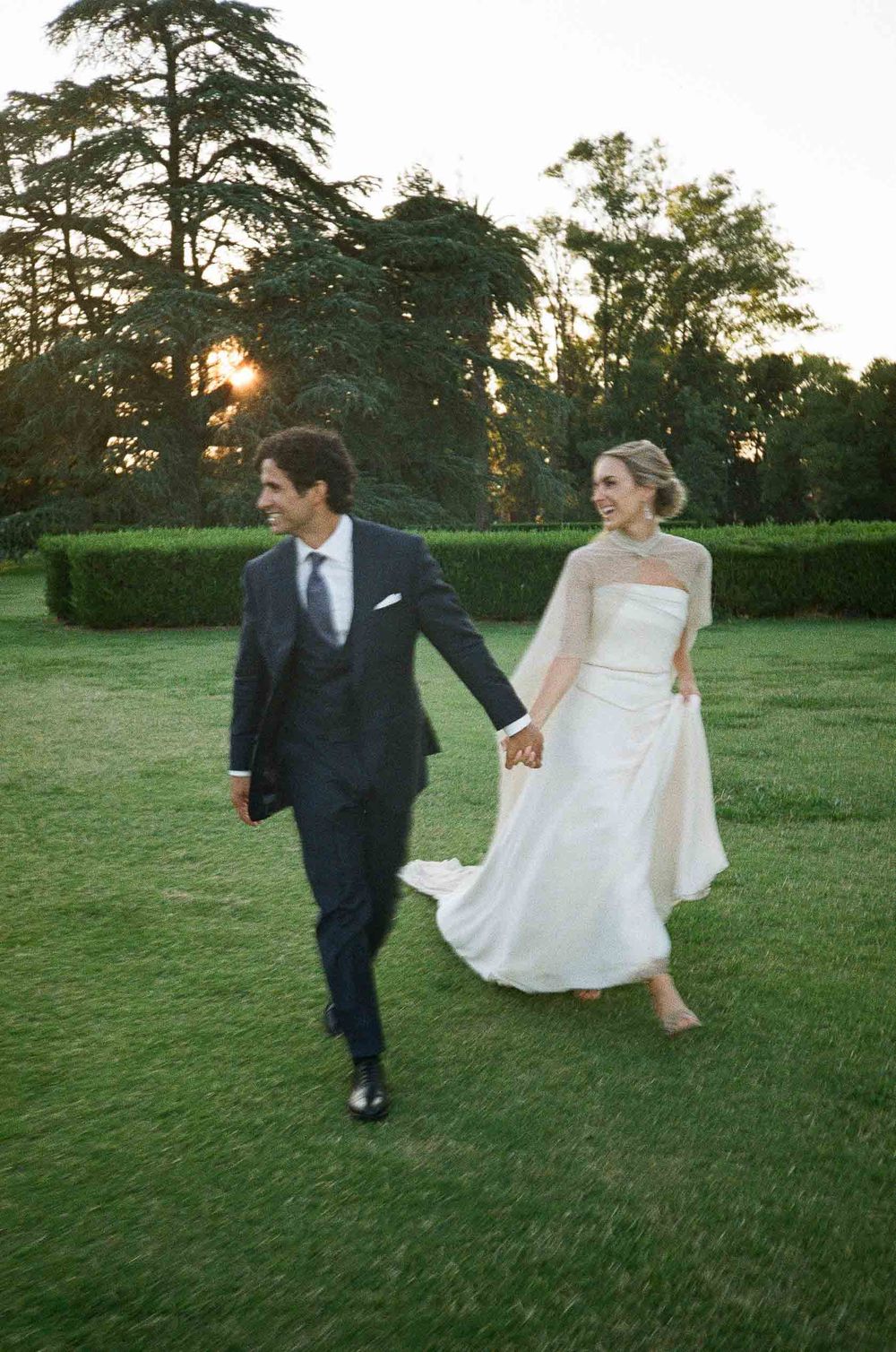 A couple in wedding attire playfully runs across a lush green lawn at sunset with tall trees in the background.