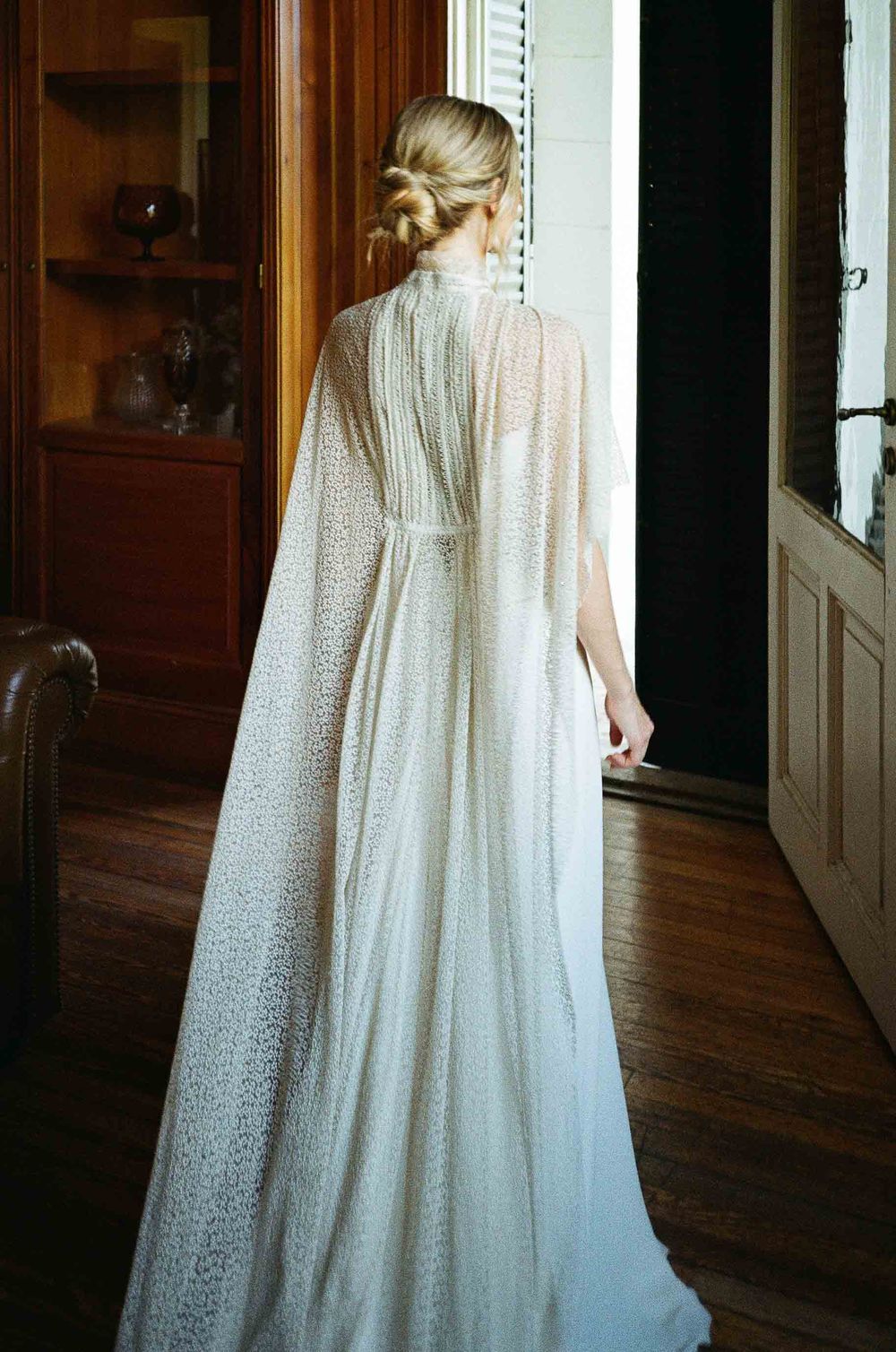 A flowing white lace wedding dress with a long train is photographed in an elegant indoor hallway.