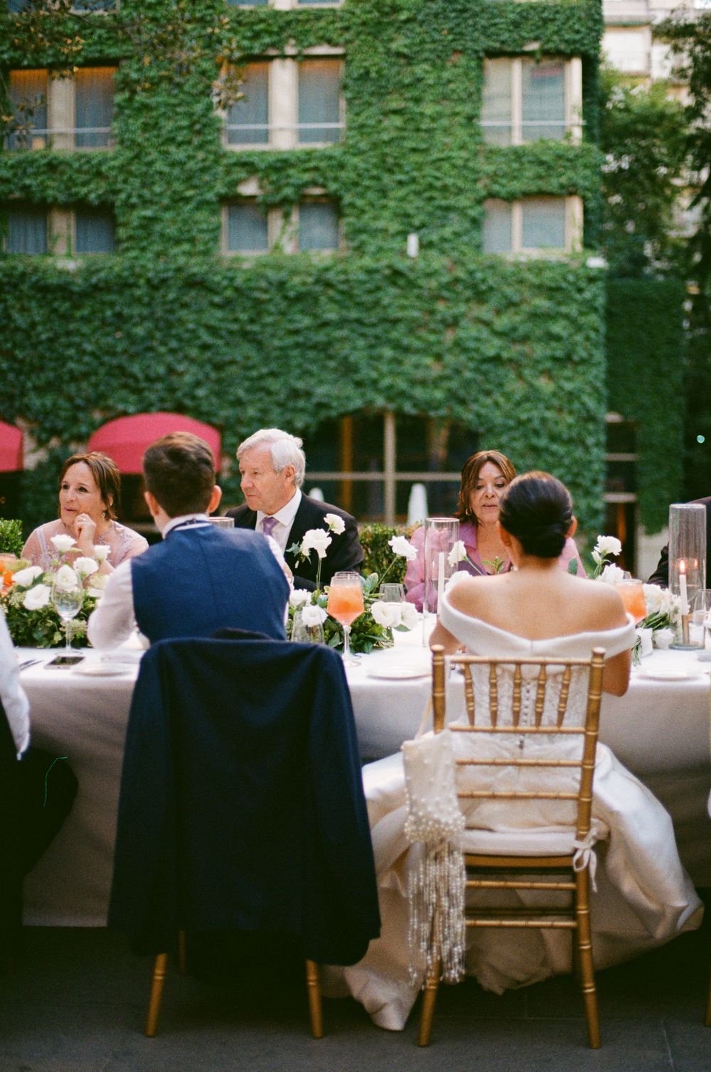 Wedding guests seated at elegant outdoor reception tables with ivy-covered walls in background.