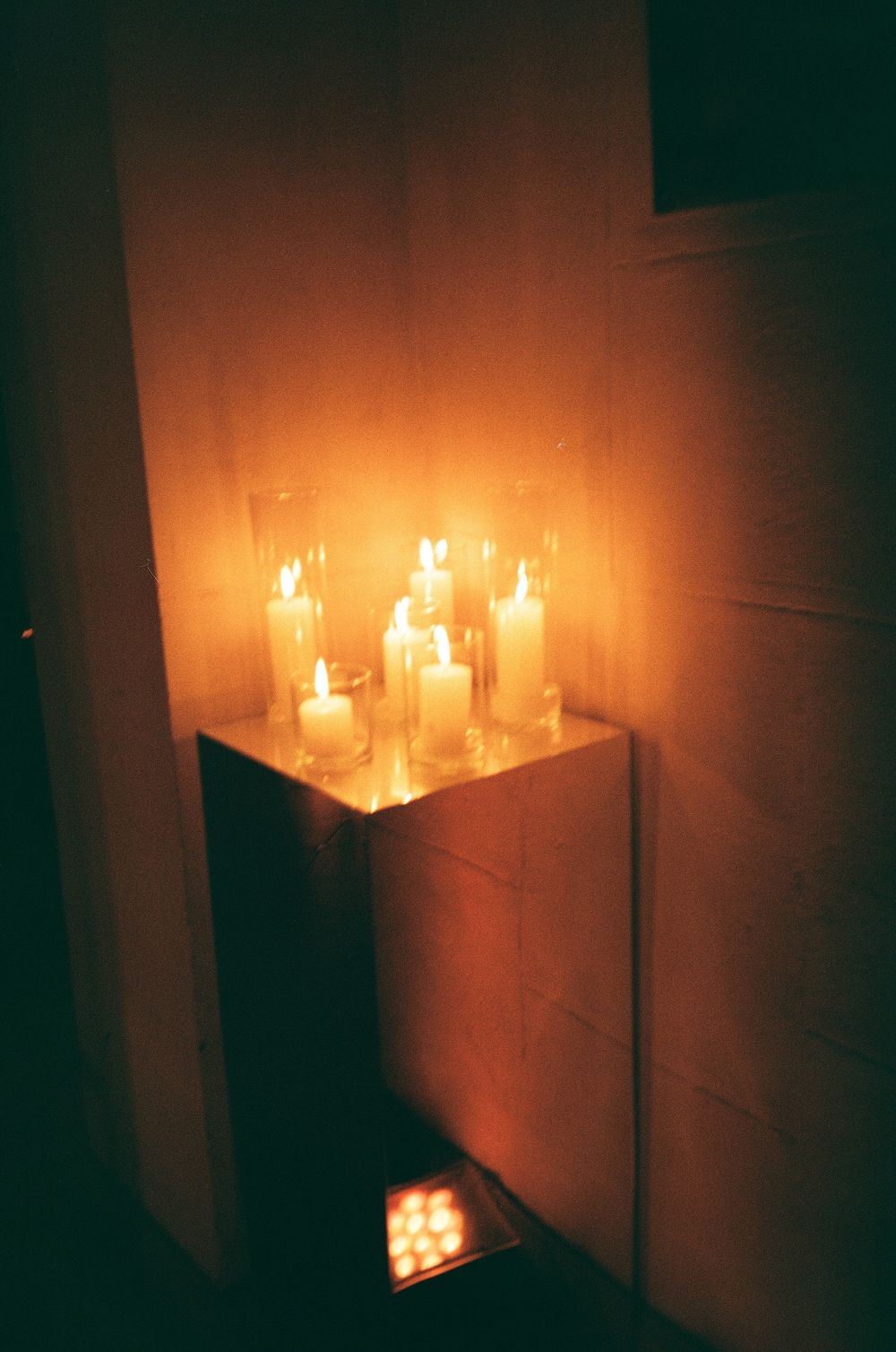 Multiple glowing candles arranged on a white shelf create a warm, atmospheric lighting effect in a dark room.