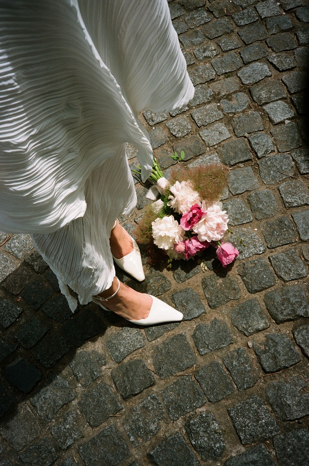 A series of photos showing white dress hem and feet walking on cobblestone pavement with pink flowers falling.