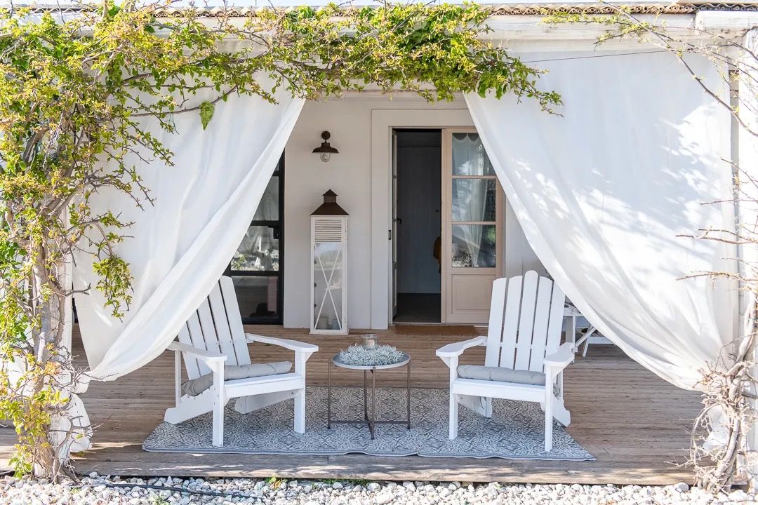 A bright white porch with draped curtains, two Adirondack chairs, and greenery creating a calm outdoor seating area.