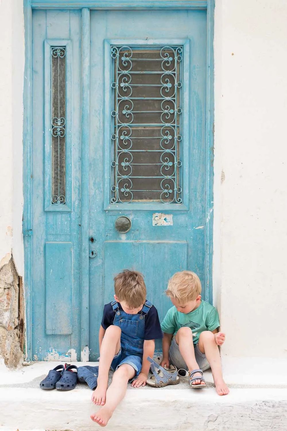 Children's feet and legs sit on a white doorstep in front of a turquoise blue door with ornate metal window grates.