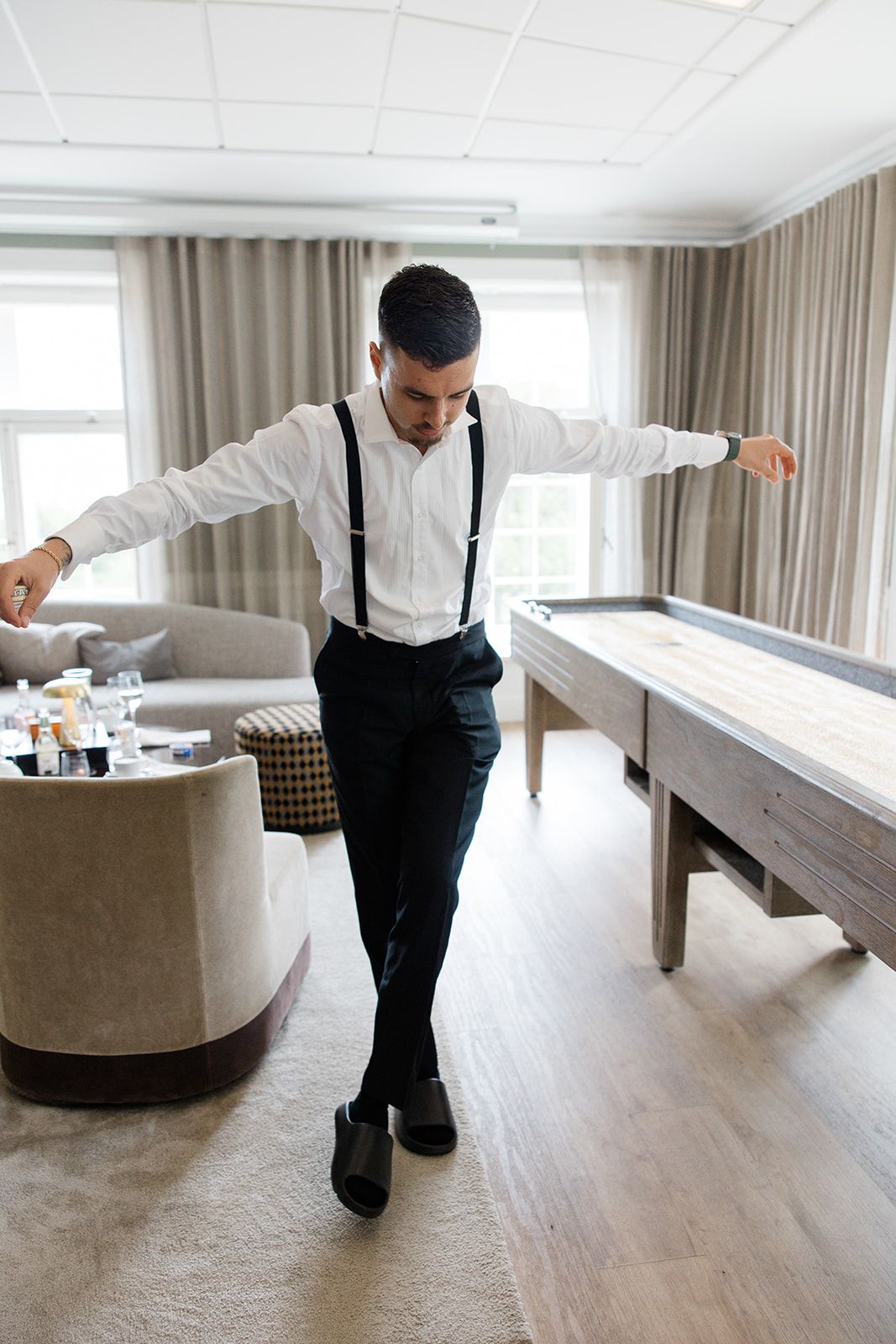 A professional cleaner in suspenders and dress shirt carefully vacuuming a beige carpet in a modern living room.