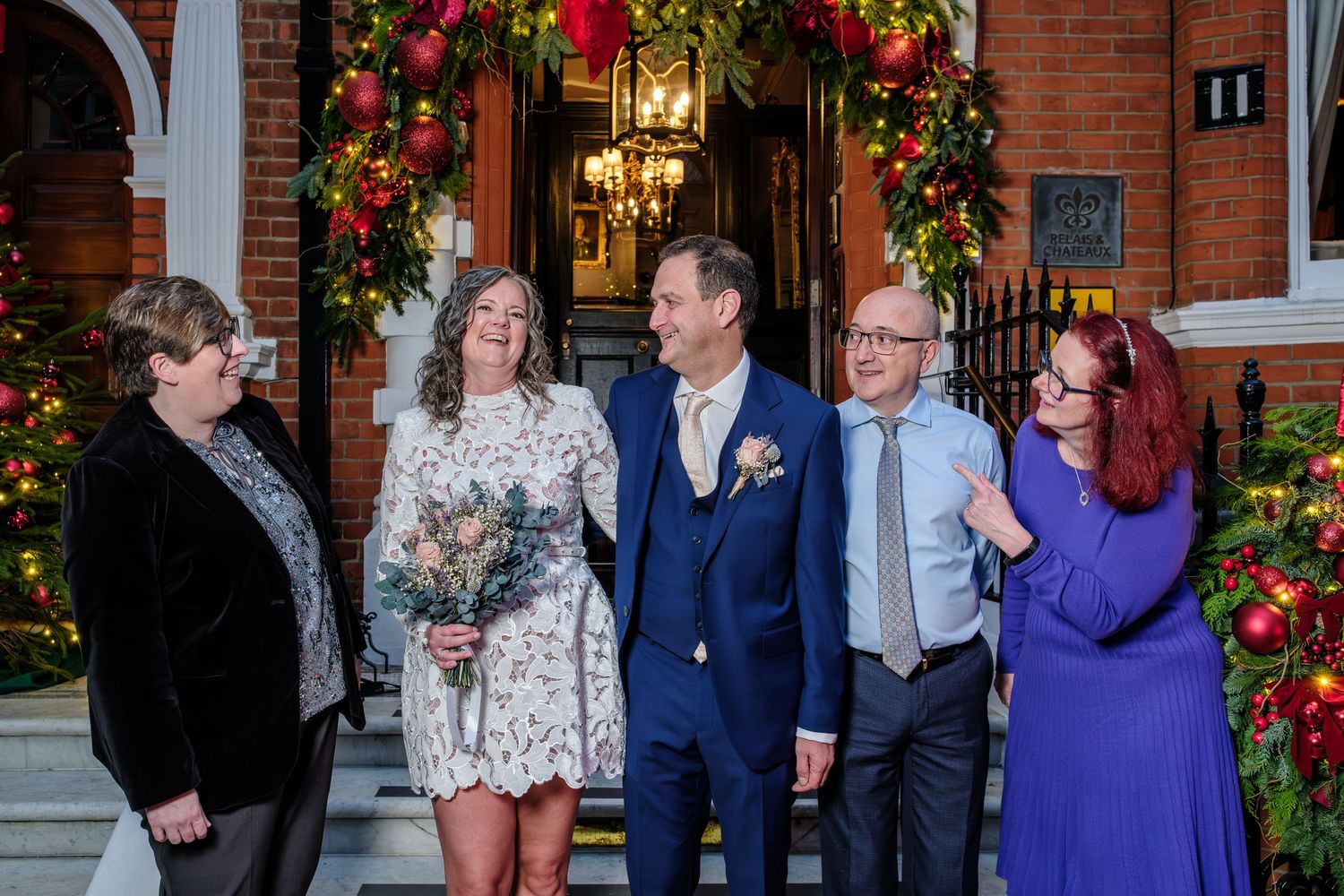 Wedding guests gather and socialize outside a brick building decorated with bright red floral arrangements.