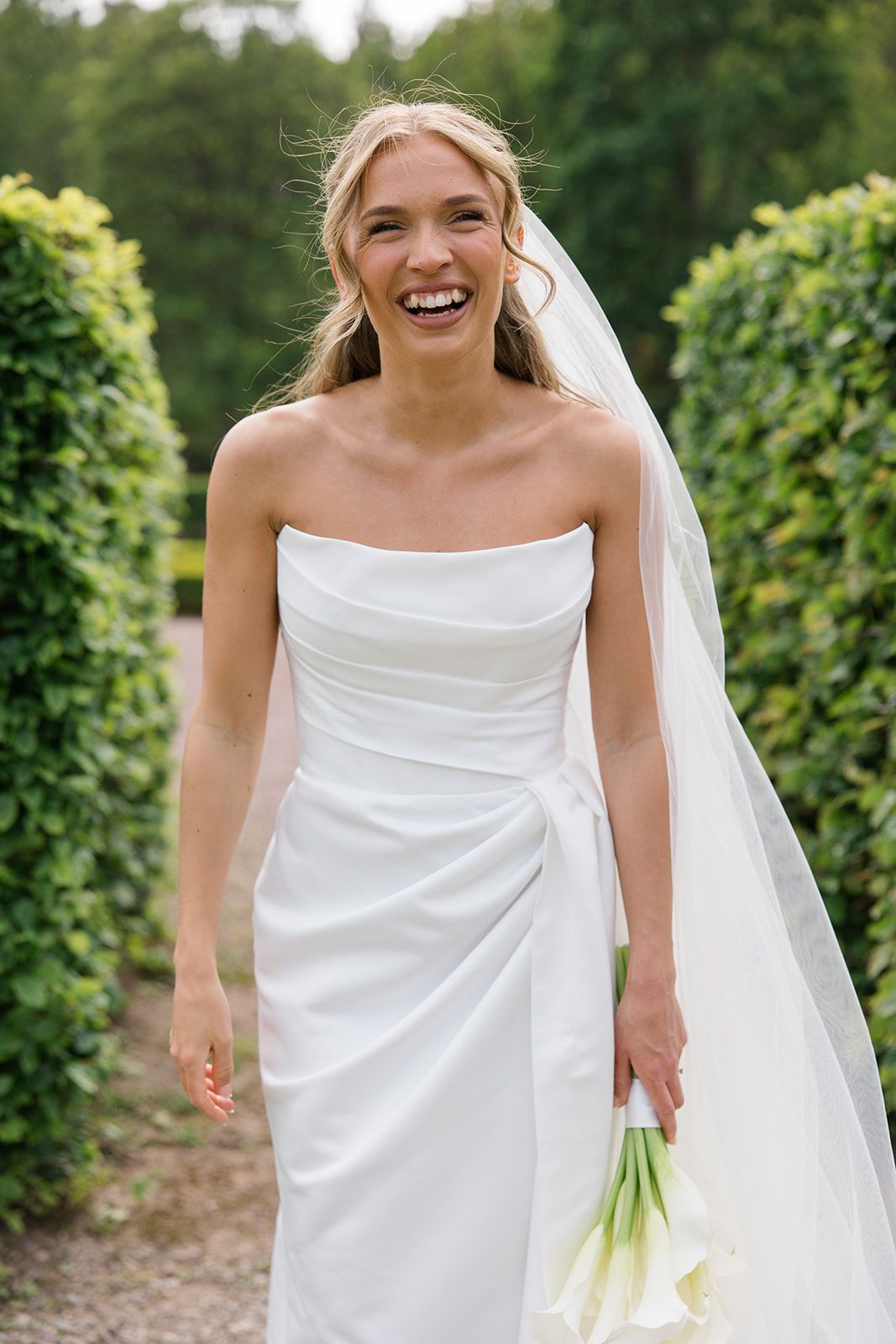 A joyful bride in a white strapless wedding dress and veil holds a bouquet in a garden setting.