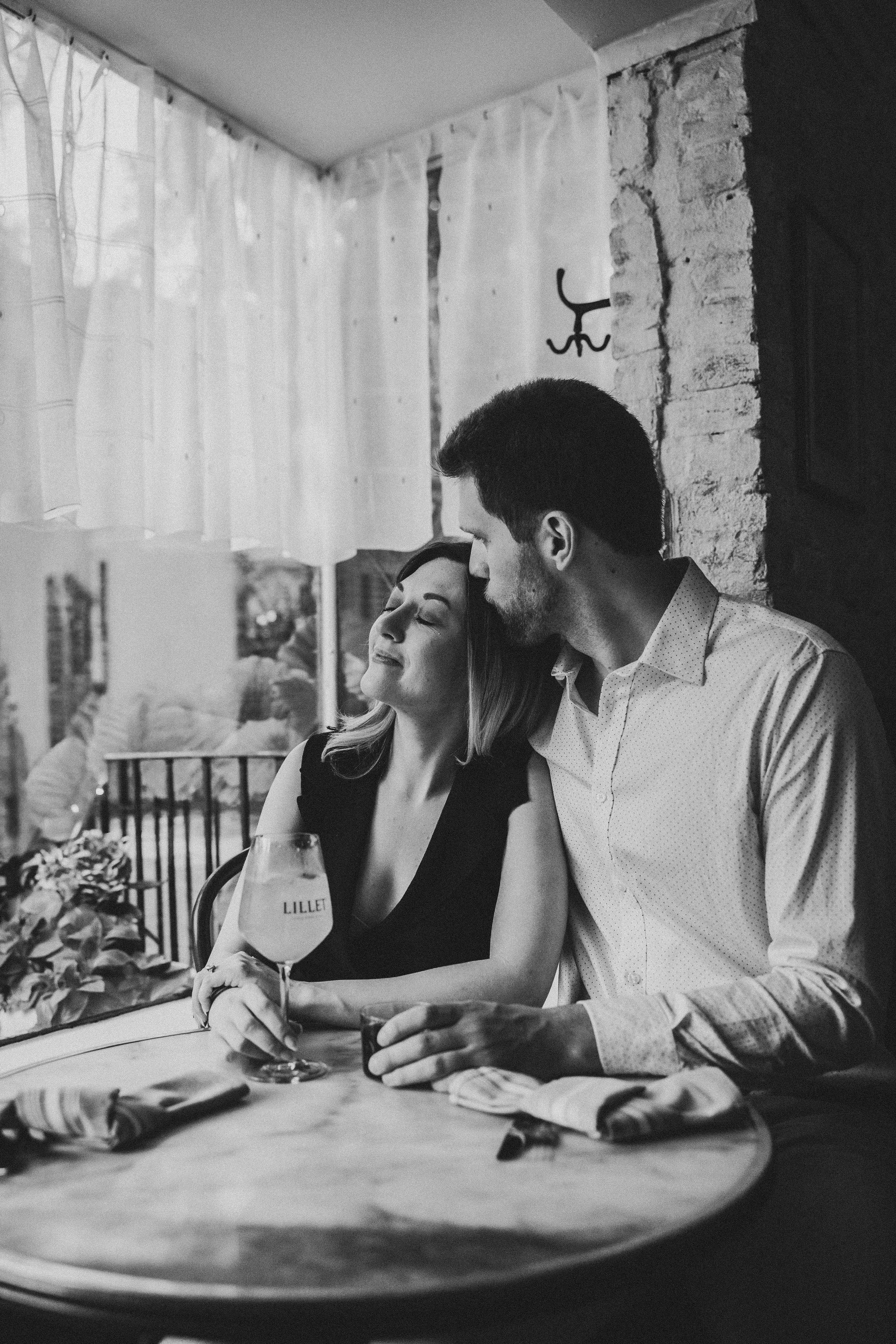 A romantic black and white photo of a couple sharing an intimate moment at a cafe table by a window.