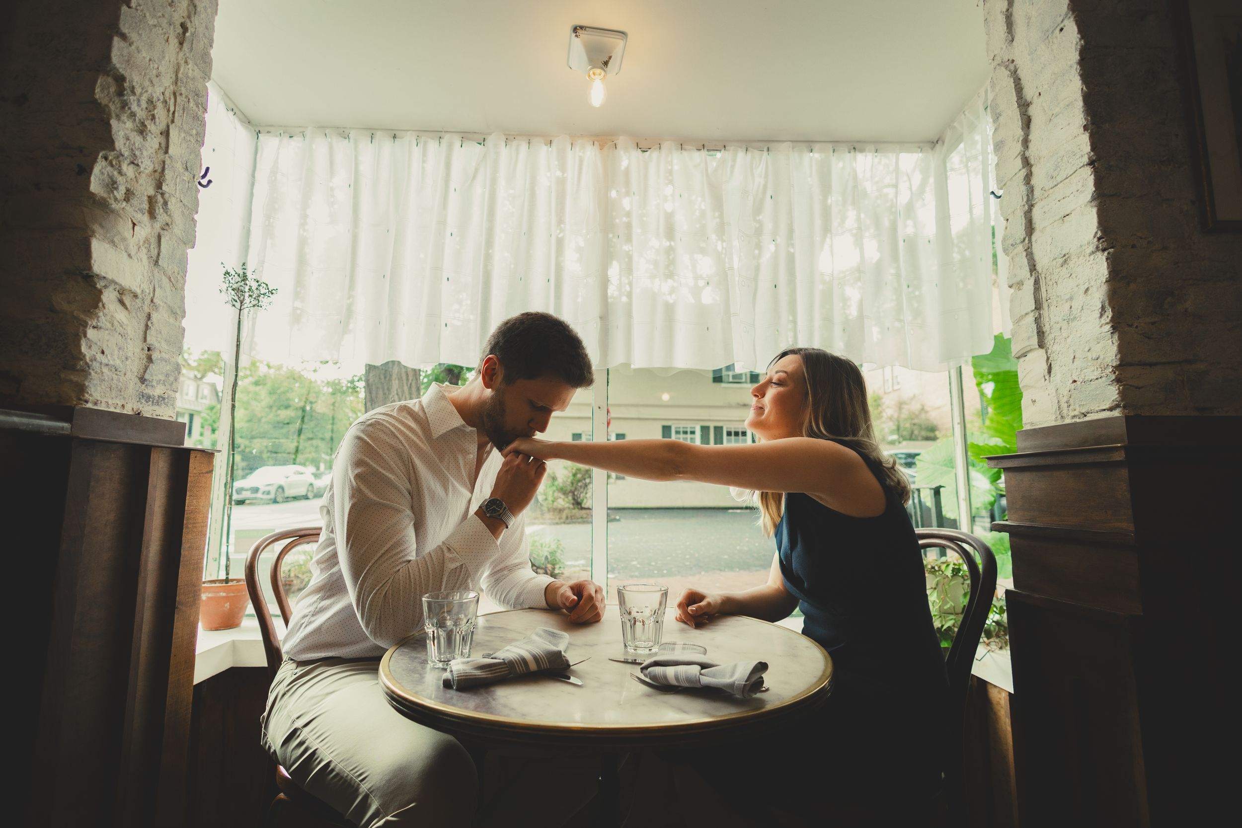 Hannah and Connor sitting by the window at La Bonne Vache in Georgetown as Connor kisses Hannah’s hand during their candid DC engagement pho