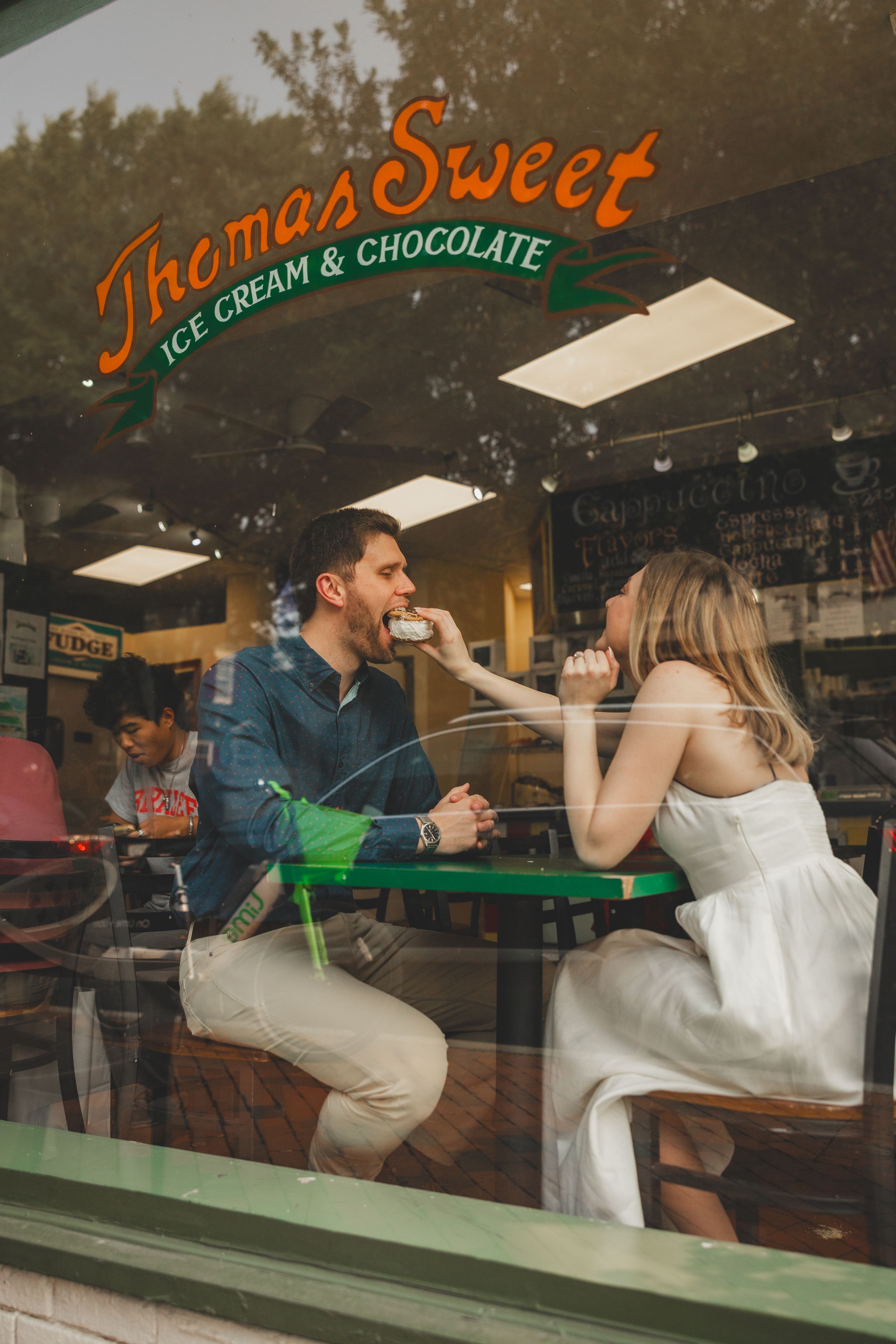 A couple shares ice cream at a retro ice cream shop counter through a window reflection.