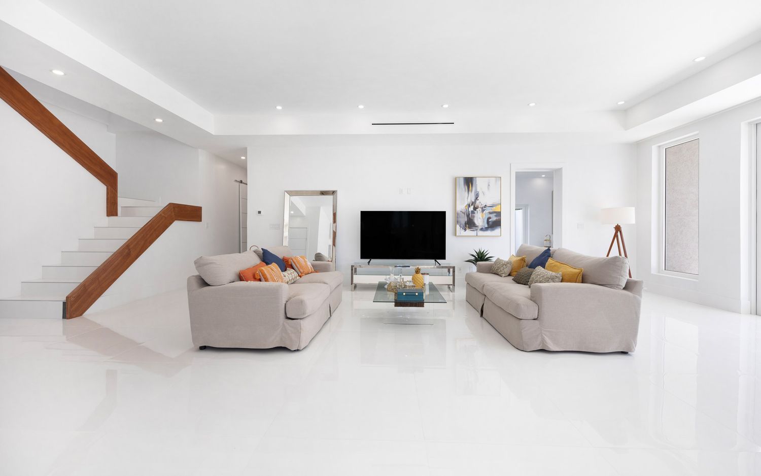 Modern white living room interior with beige sofas, wooden staircase, and wall-mounted TV in minimalist design.