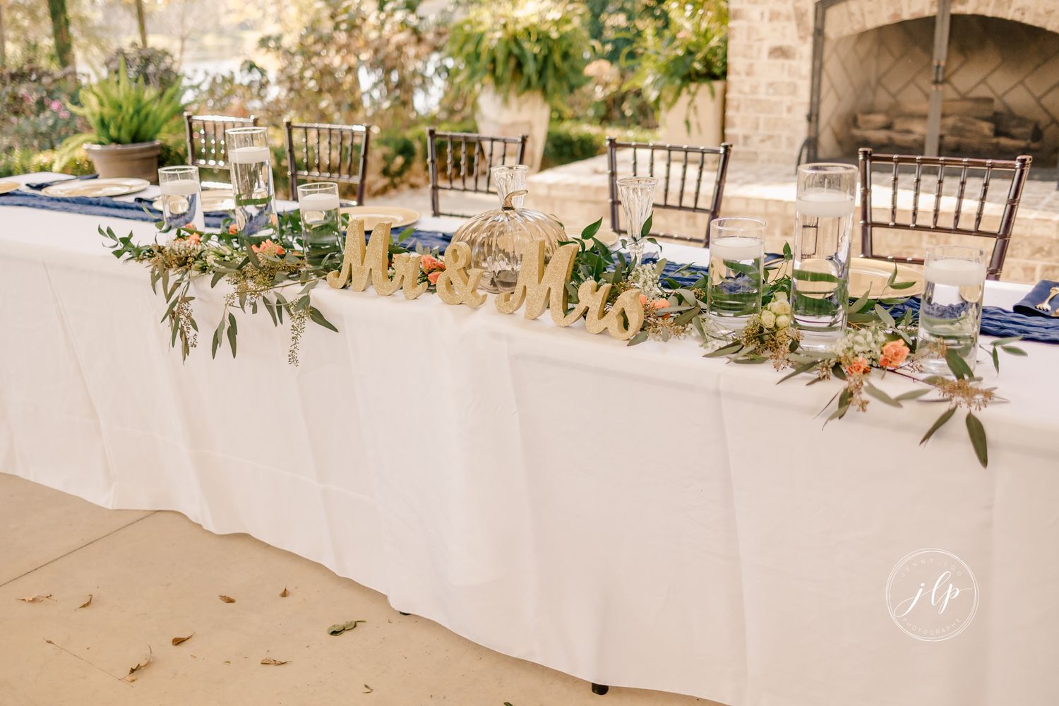A rustic wedding reception table setup with white linens and wooden chairs in an outdoor venue.