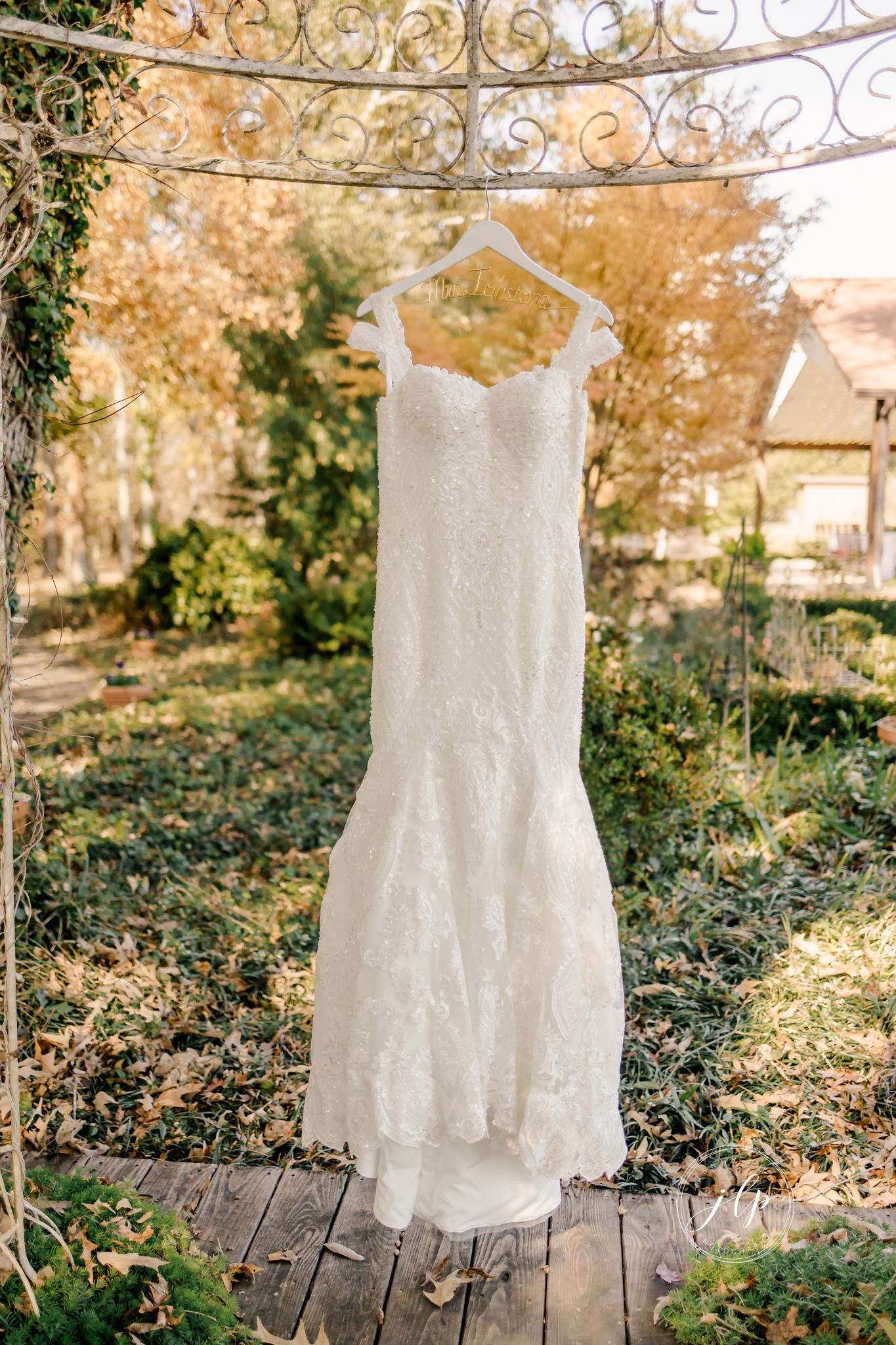 White lace wedding dress hanging from ornate iron gazebo with autumn foliage in background.