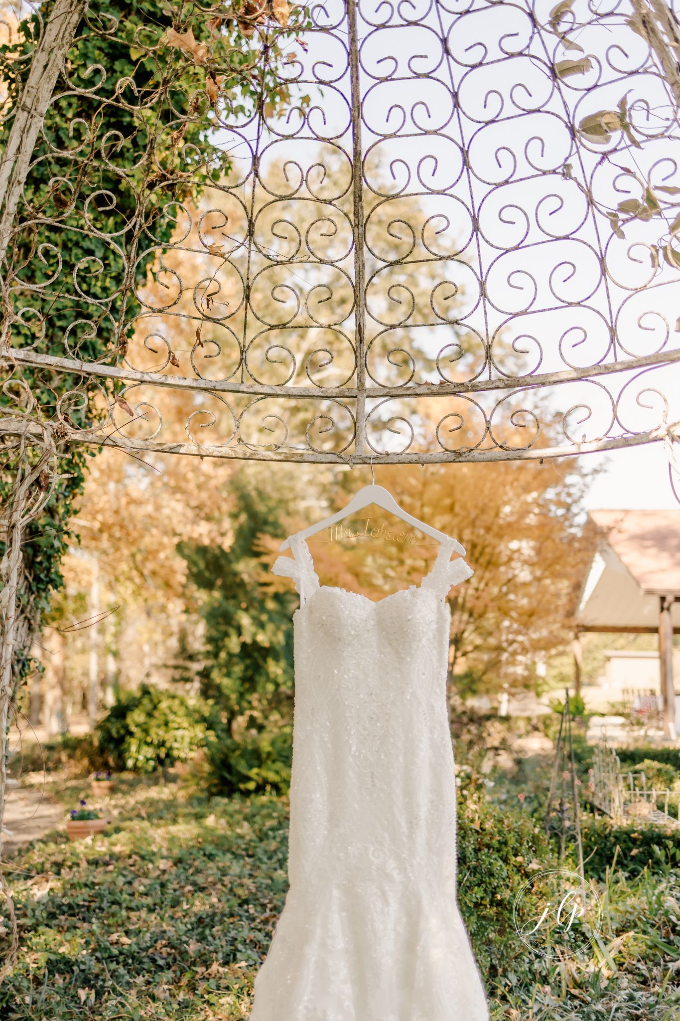 An elegant white wedding dress hanging from an ornate metal gazebo in a garden setting.