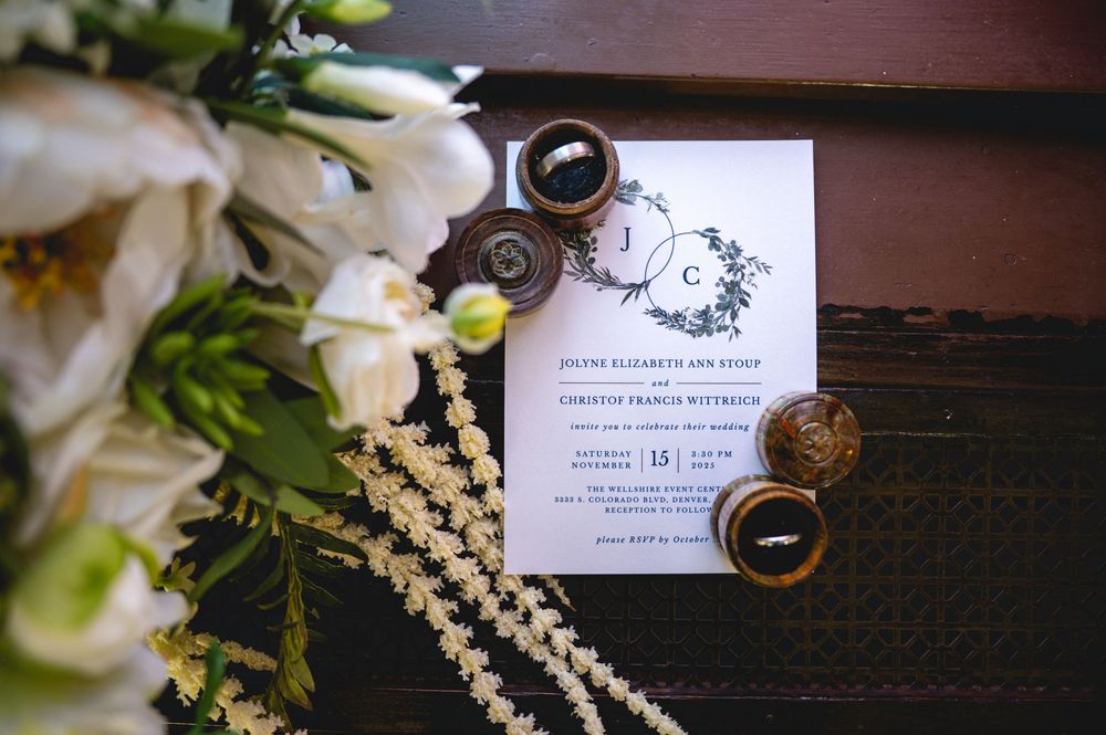 Wedding rings and invitation details artistically arranged with white flowers and greenery on a wooden surface.