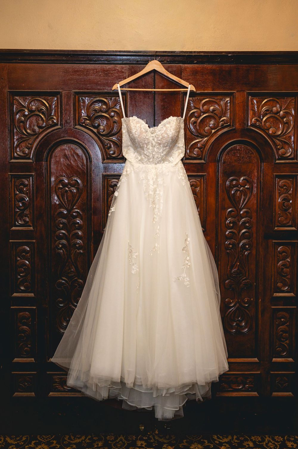 White tulle wedding dress with lace bodice hangs elegantly against ornate carved wooden paneling.