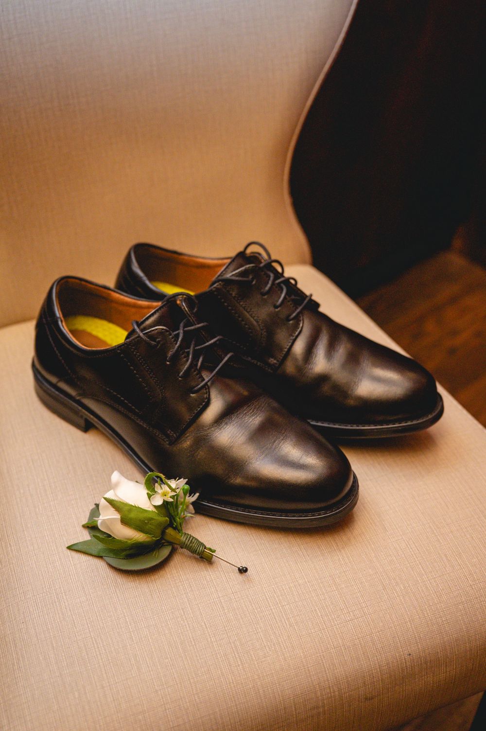 A pair of classic black leather dress shoes with laces sits on a beige surface next to a small green boutonniere.