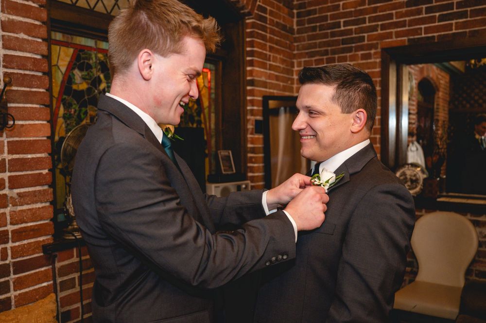 Two people in suits share a laugh while standing in front of a brick wall in an office or business setting.