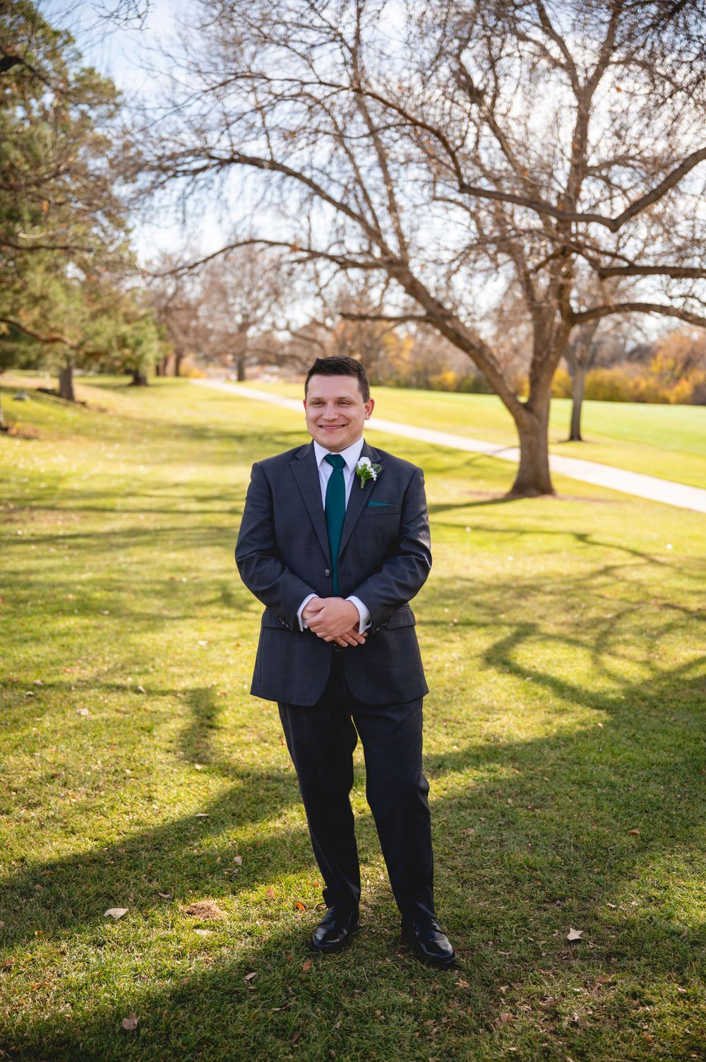 A person in a dark suit and teal tie poses outdoors on grassy parkland with trees on a sunny autumn day.
