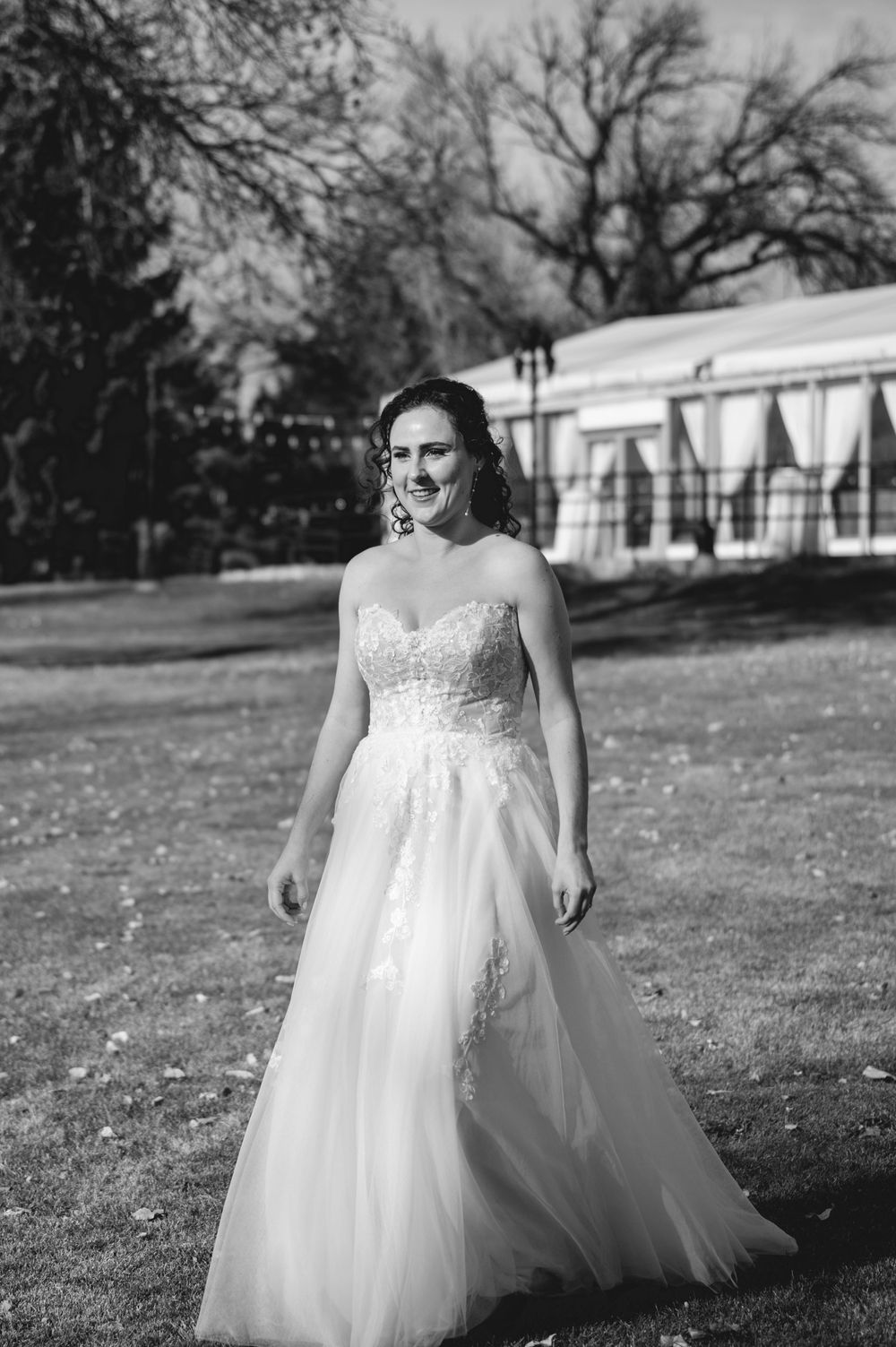 A bride in a flowing white lace and tulle ballgown poses outdoors on a grassy lawn near white buildings.