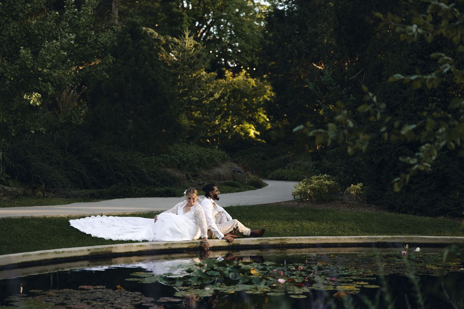 A couple in wedding attire lies on a wooden dock surrounded by lily pads and trees at sunset.