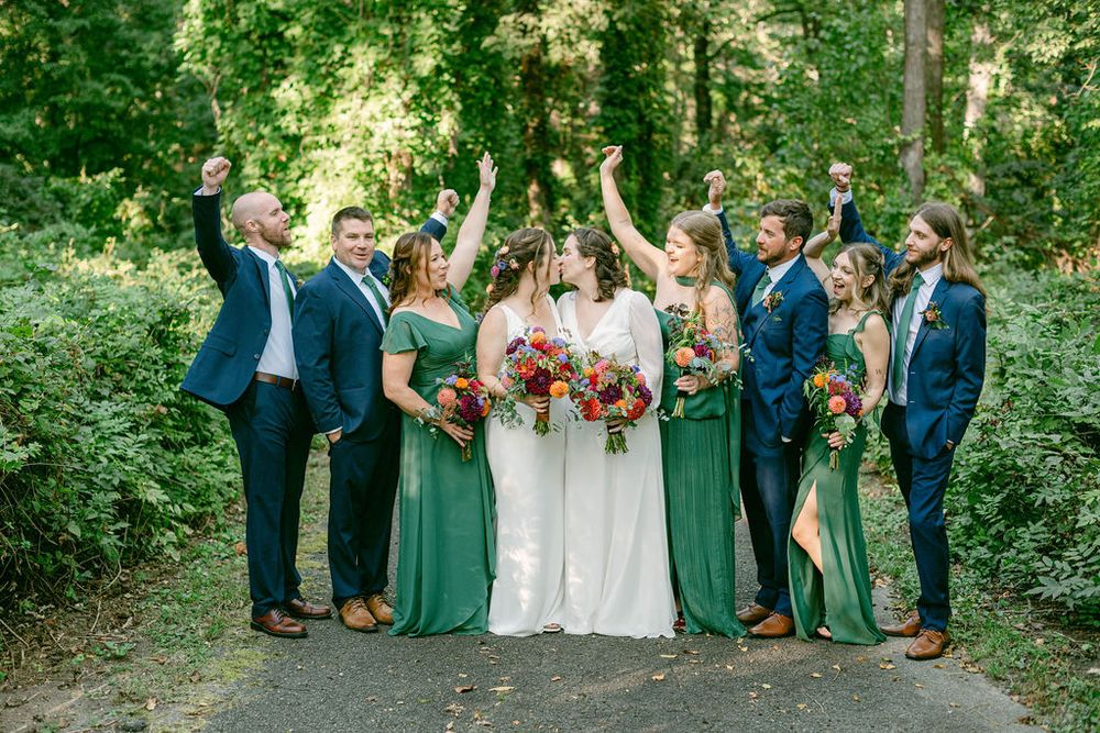 Wedding party in navy and emerald green attire celebrates joyfully on a forest path with vibrant bouquets.