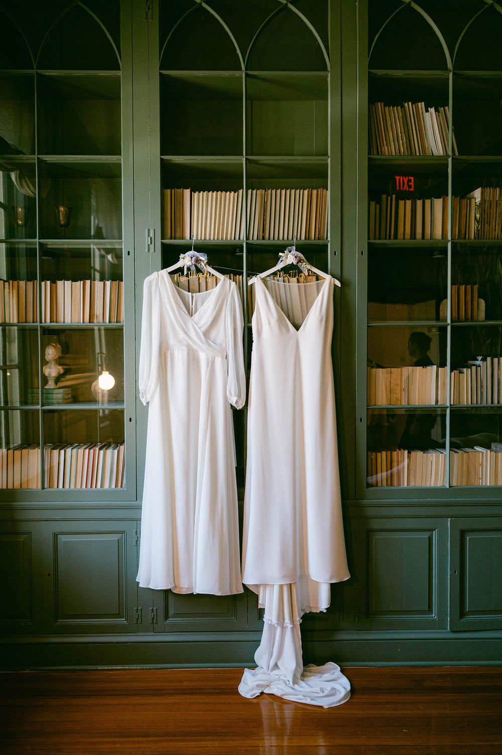 White bridesmaid dresses hanging on hangers against dark wooden bookshelves in a library setting.