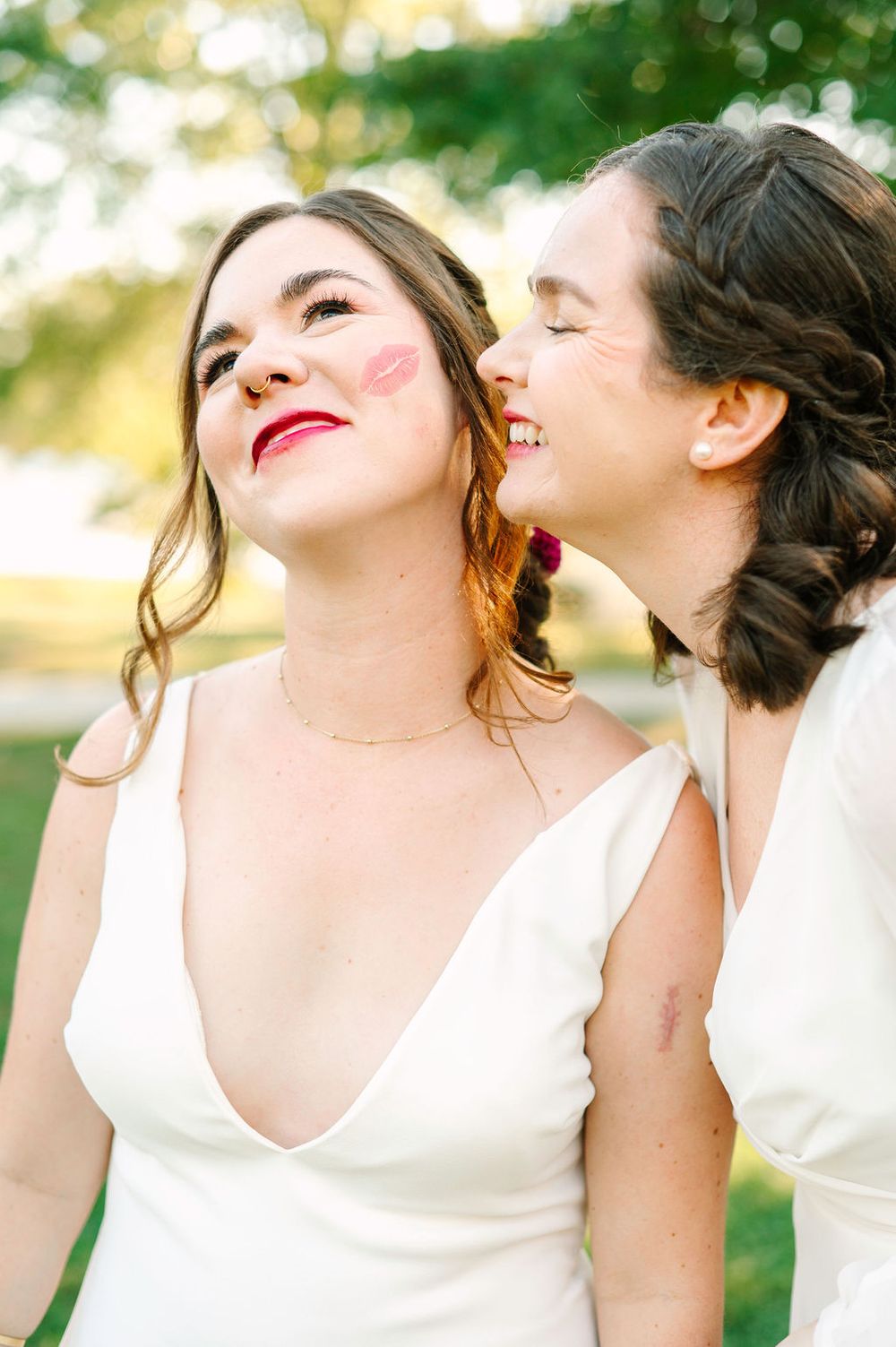 Two people in white dresses sharing a joyful candid moment outdoors with natural sunlit bokeh.