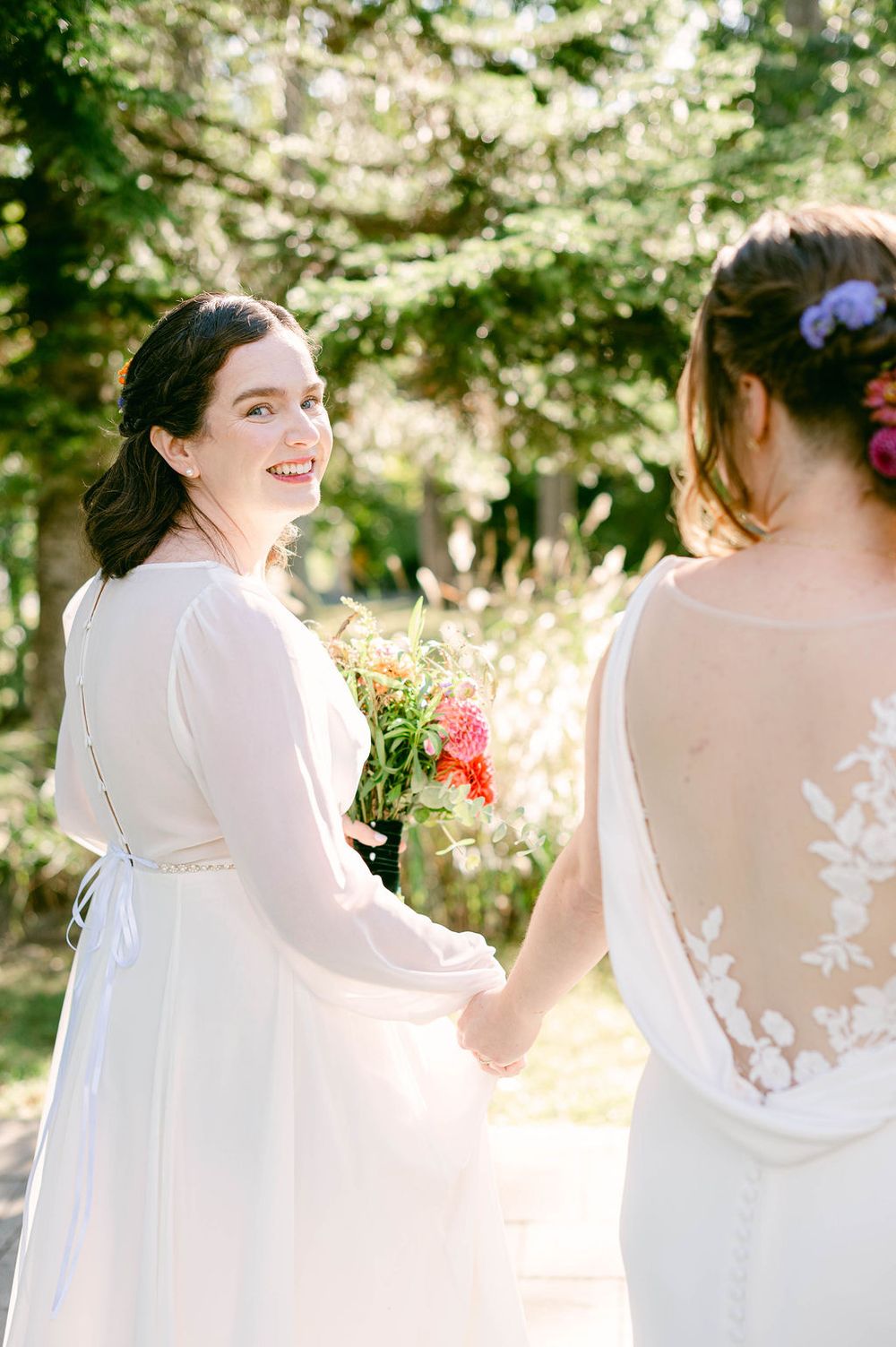 White-dressed figures holding bouquets in sunlit outdoor garden setting.