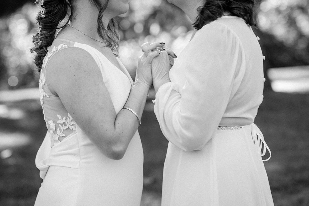 Black and white photograph showing two people in white dresses holding hands in an intimate outdoor moment.