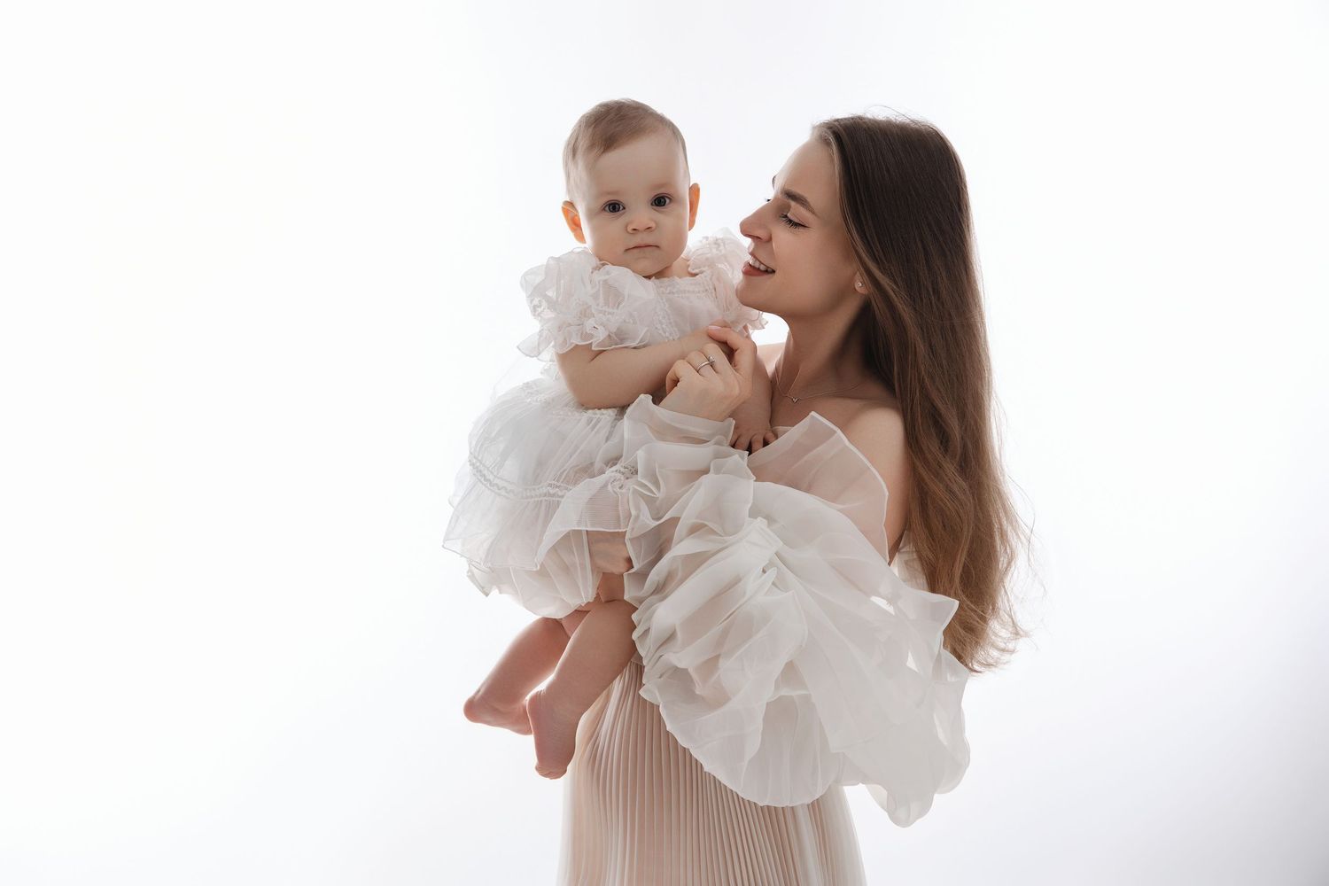 A parent in white clothing lifts and plays with a baby wearing a white dress against a bright background.