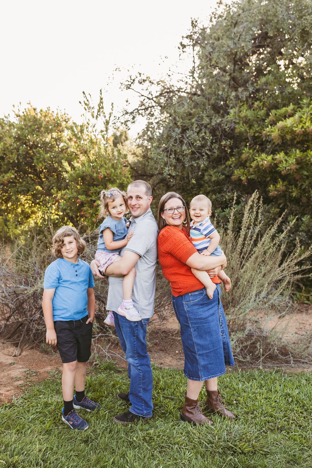 A family poses together outdoors in coordinating blue and red outfits against a natural backdrop during golden hour.