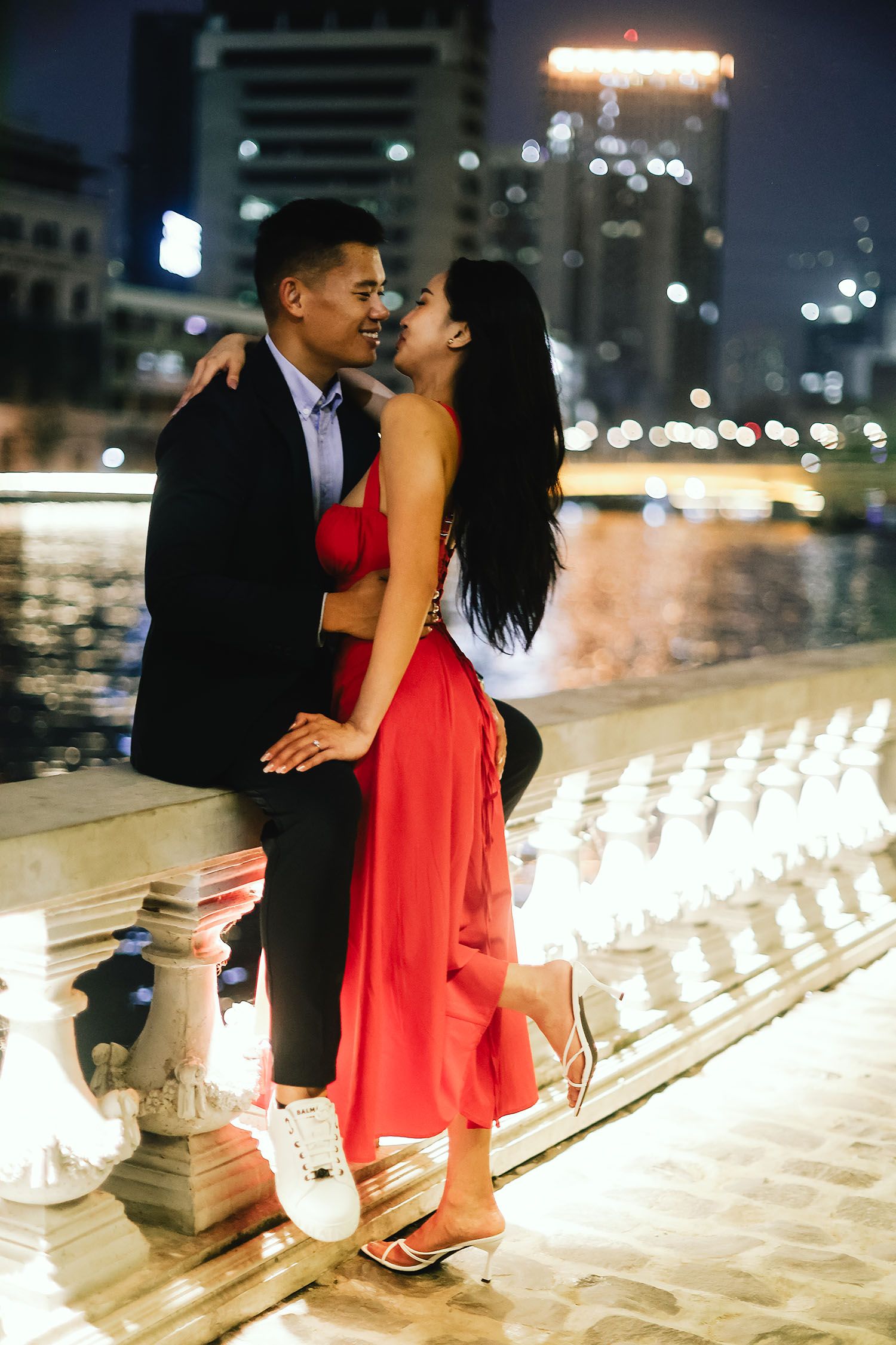 Romantic couple in evening attire embraces on illuminated steps overlooking city lights and waterfront.