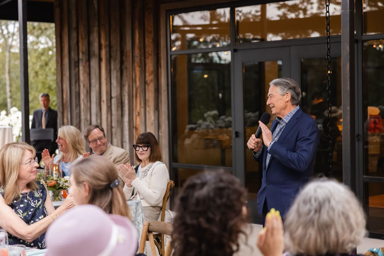 A speaker addresses a seated audience during an outdoor event at a modern venue with wooden architectural details.