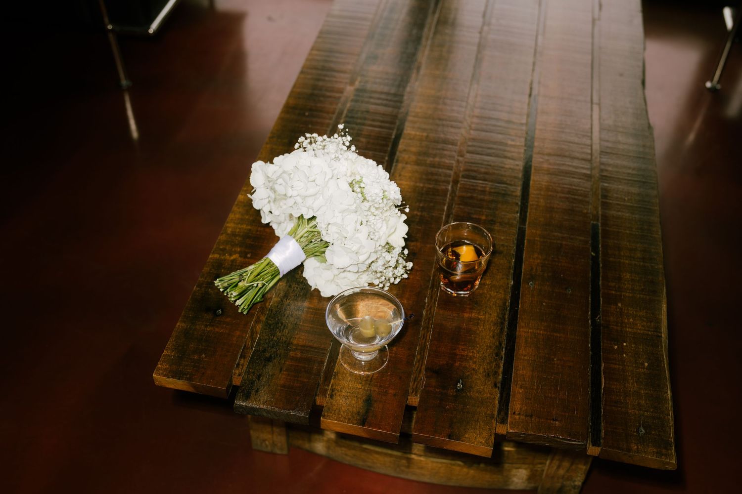 A white floral bouquet in a glass vase sits on a dark wooden table with a small candle holder nearby.