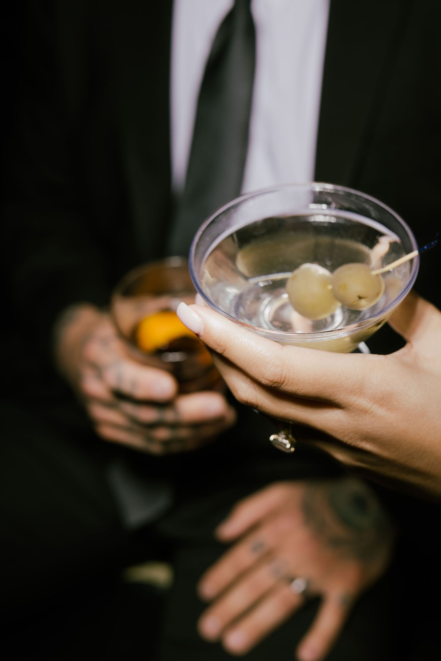 Multiple hands hold coupe cocktail glasses with clear drinks and garnishes in a dimly lit setting.