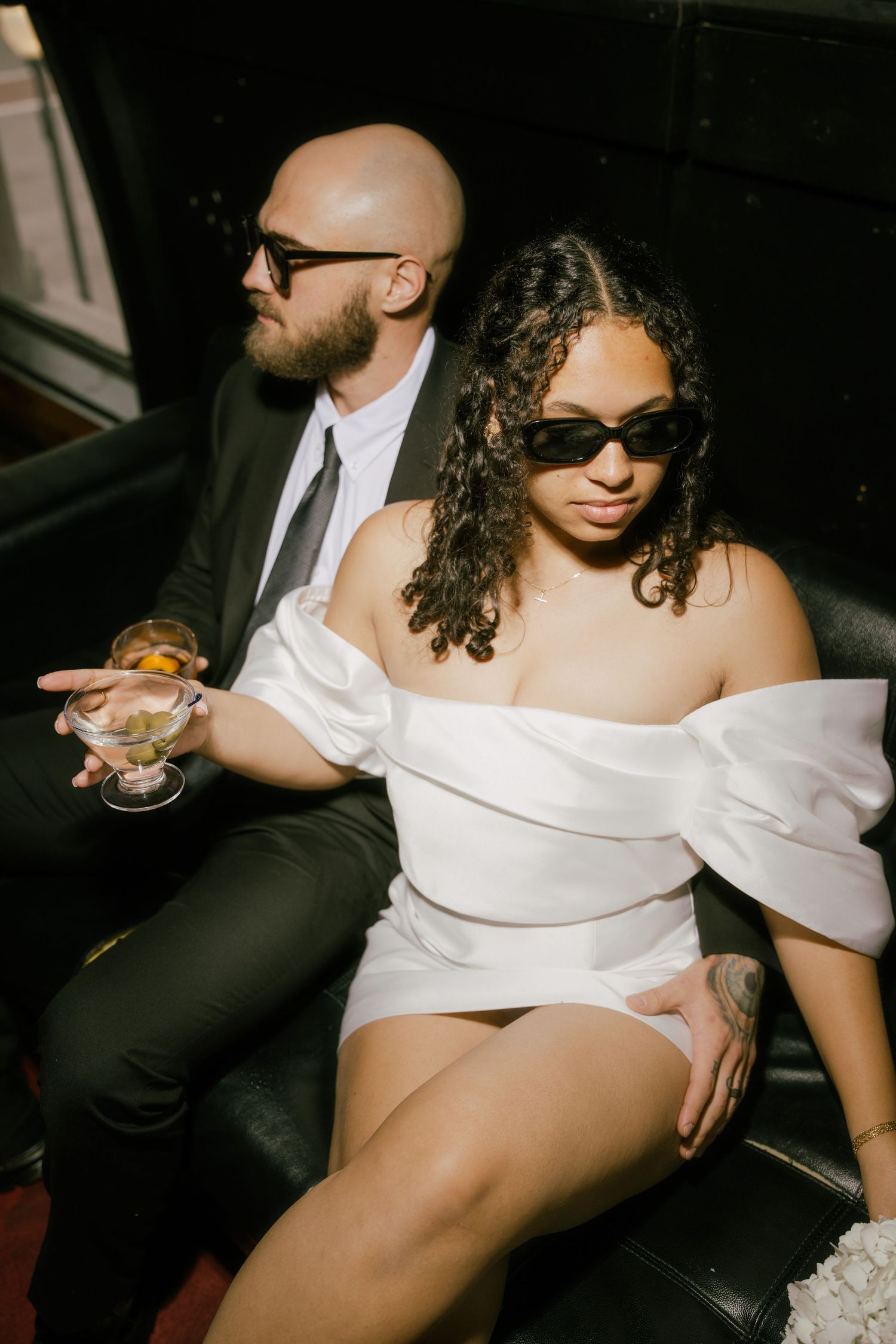 A couple lounges intimately on a dark leather couch with wine glasses in a dimly lit setting.